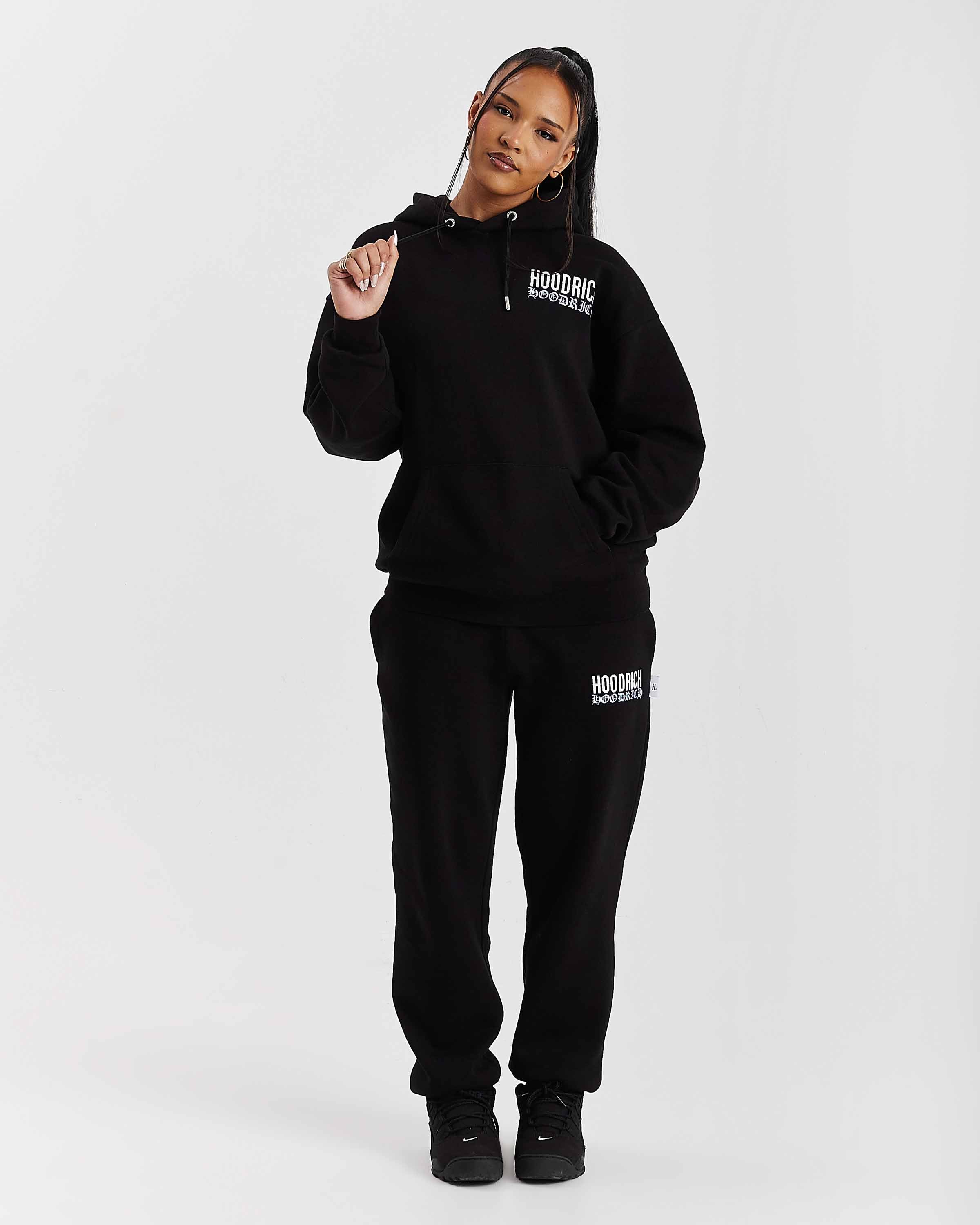 Person wearing a black hoodie and pants with 'Hoodrich' branding on a white background