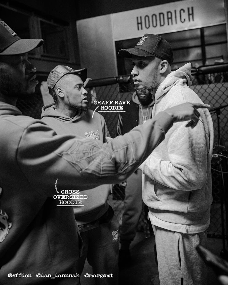A black and white streetwear photograph featuring three men in a candid urban setting. One man on the right wears a light-colored hoodie and matching sweatpants, while another points toward him.
