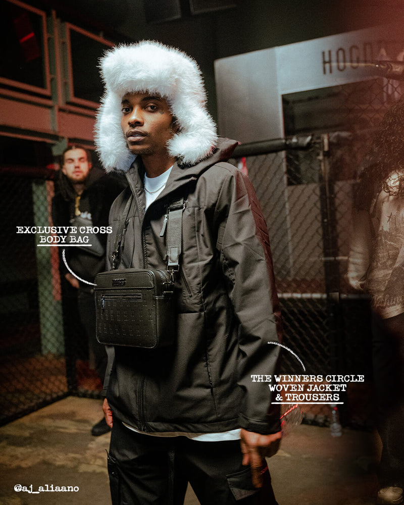 A man wearing a black crossbody bag with a white faux-fur trimmed hat and a dark jacket, standing in a gritty urban setting. Text overlays mention 'EXCLUSIVE CROSS BODY BAG' and 'THE WINNERS CIRCLE WOVEN JACKET & TROUSERS'.