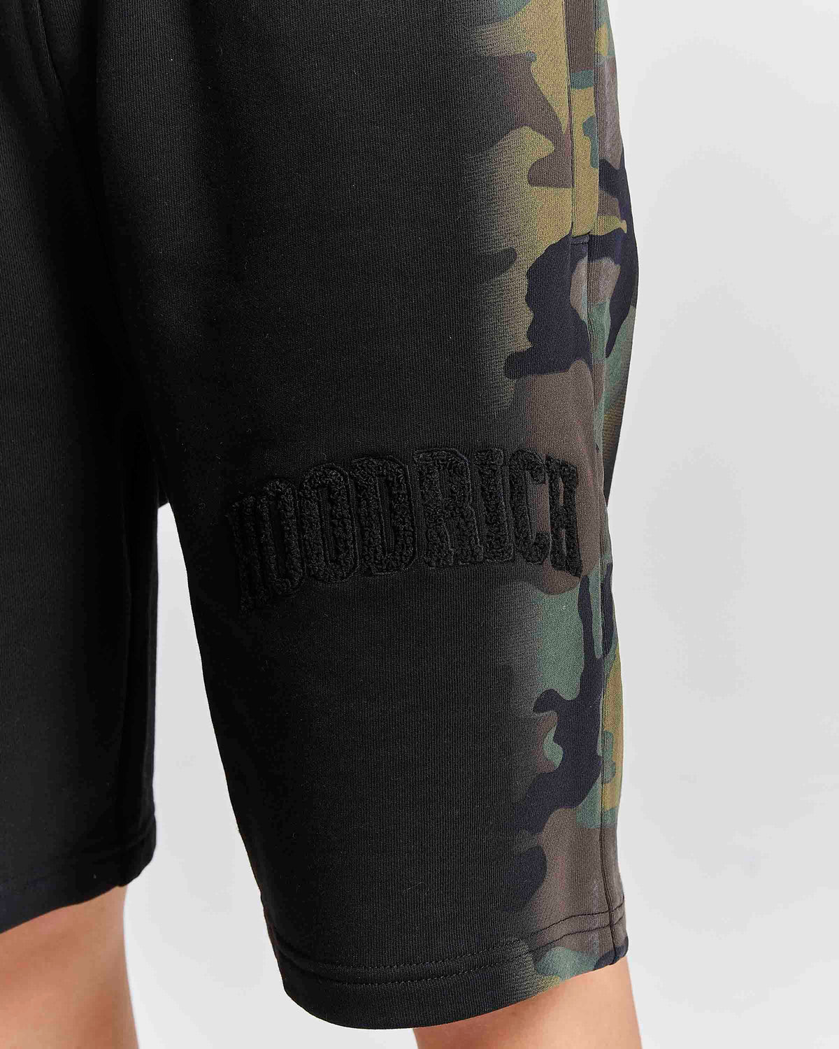 Close-up of a person wearing black shorts with a camouflage print down the side and a black embroidered 'HOODRICH' logo patch on the thigh, against a light gray background.