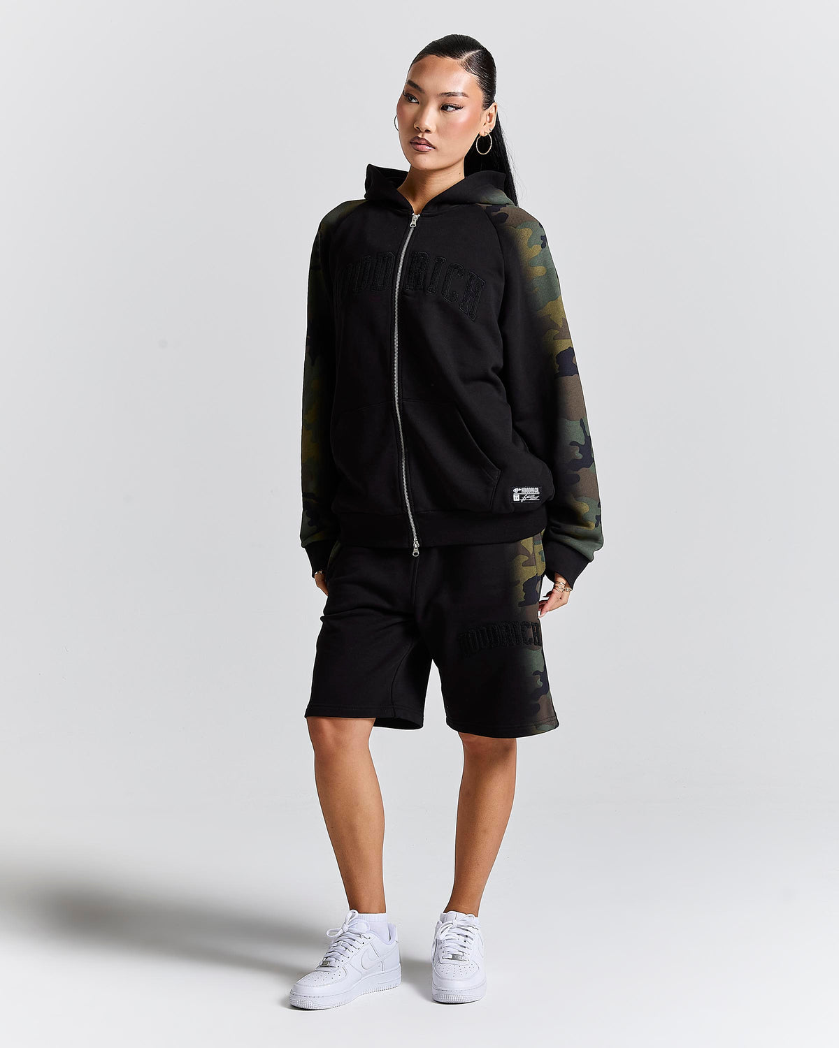 A model wearing a black hOODRICH zip-up hoodie with green camouflage sleeves, matching black Puma shorts, and white sneakers, standing against a light gray studio background.