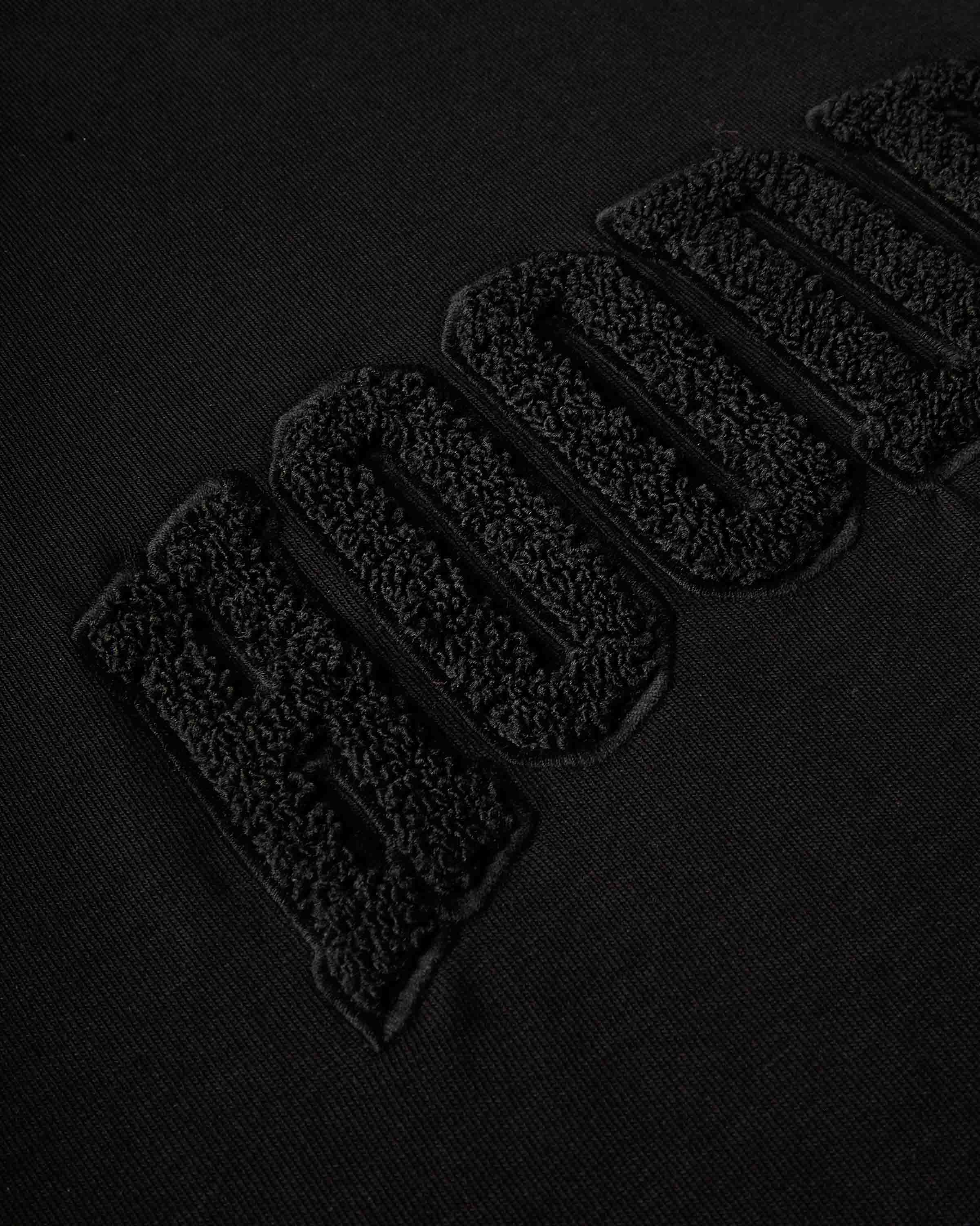 Close-up of a black textured fabric with large raised embroidered letters reading 'HOODRICH' in black-on-black thread.