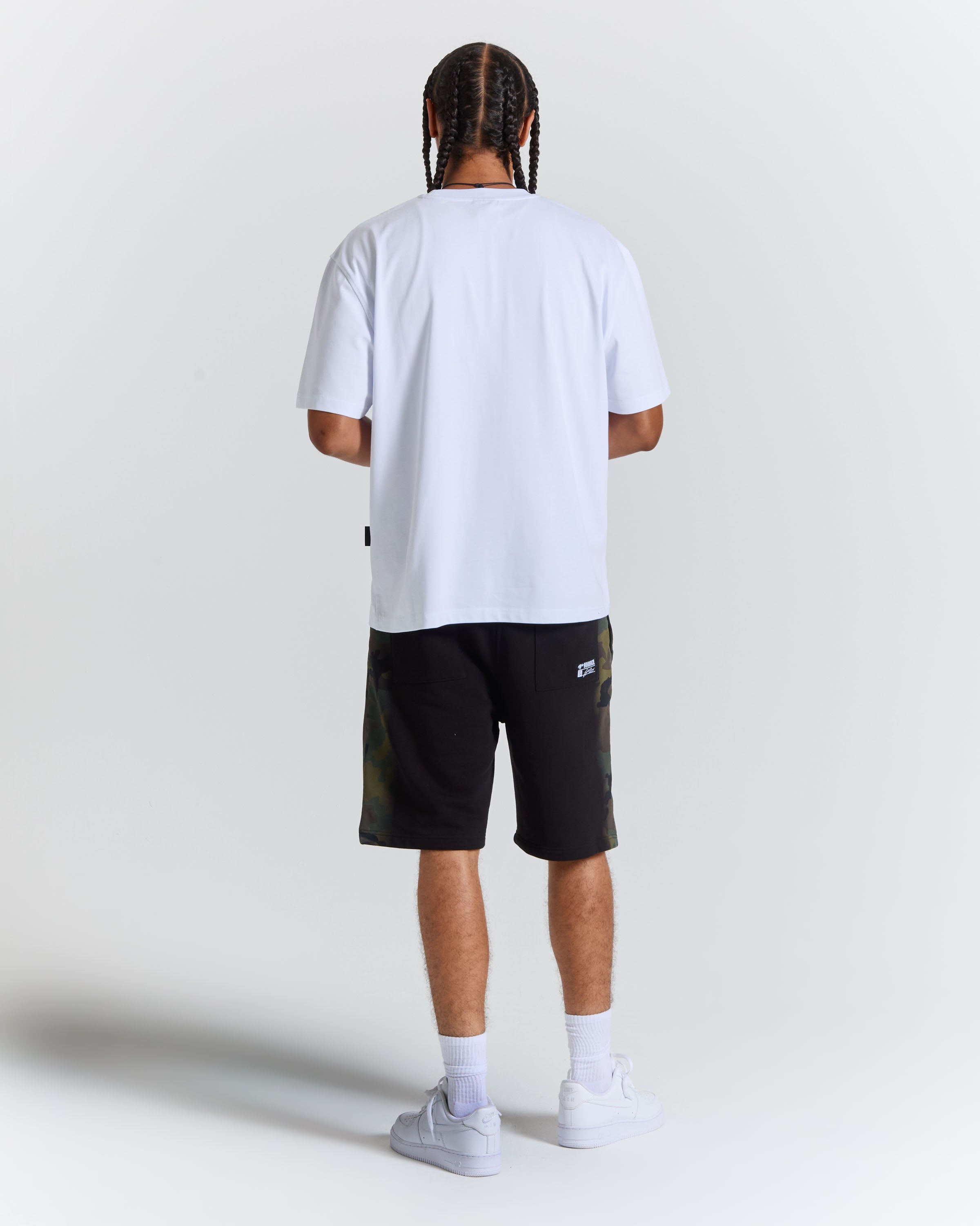 A man viewed from behind wearing a white t-shirt, black shorts with green camouflage side panels, white socks, and white sneakers, standing against a light gray studio background.