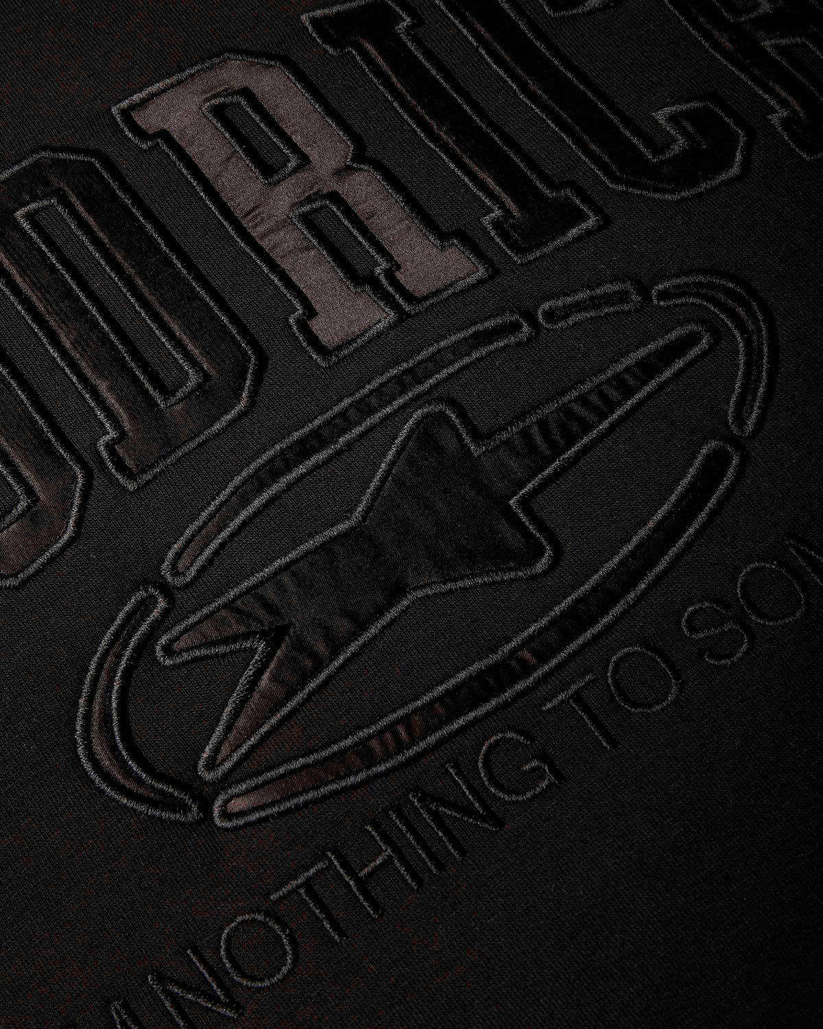 Close-up of a black fabric surface with large tonal embroidered lettering and a star-like emblem, showing partial text on HOODRICH.