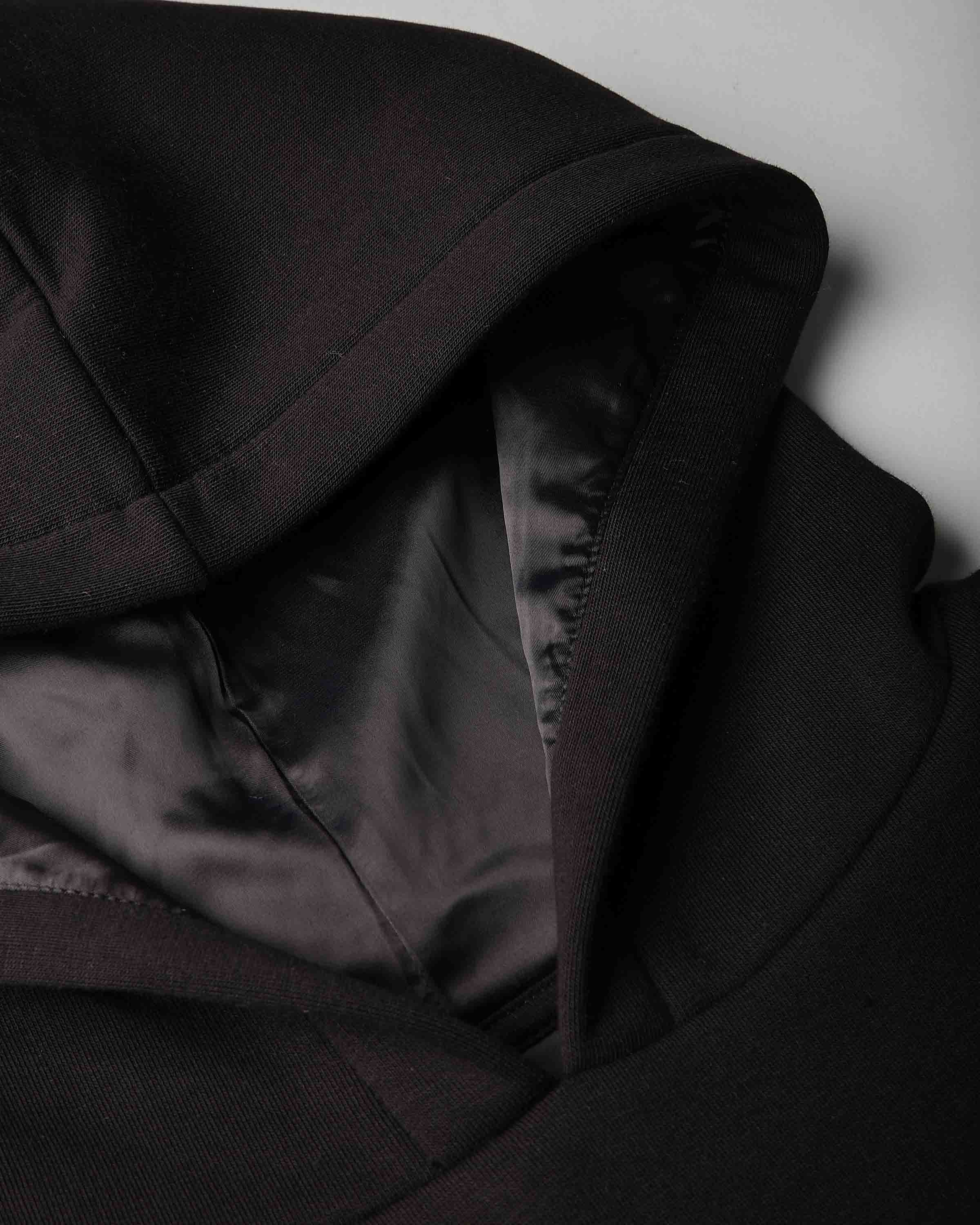 Close-up detail of a black hooded jacket or coat with a satin-like black interior lining, shown against a light gray background.