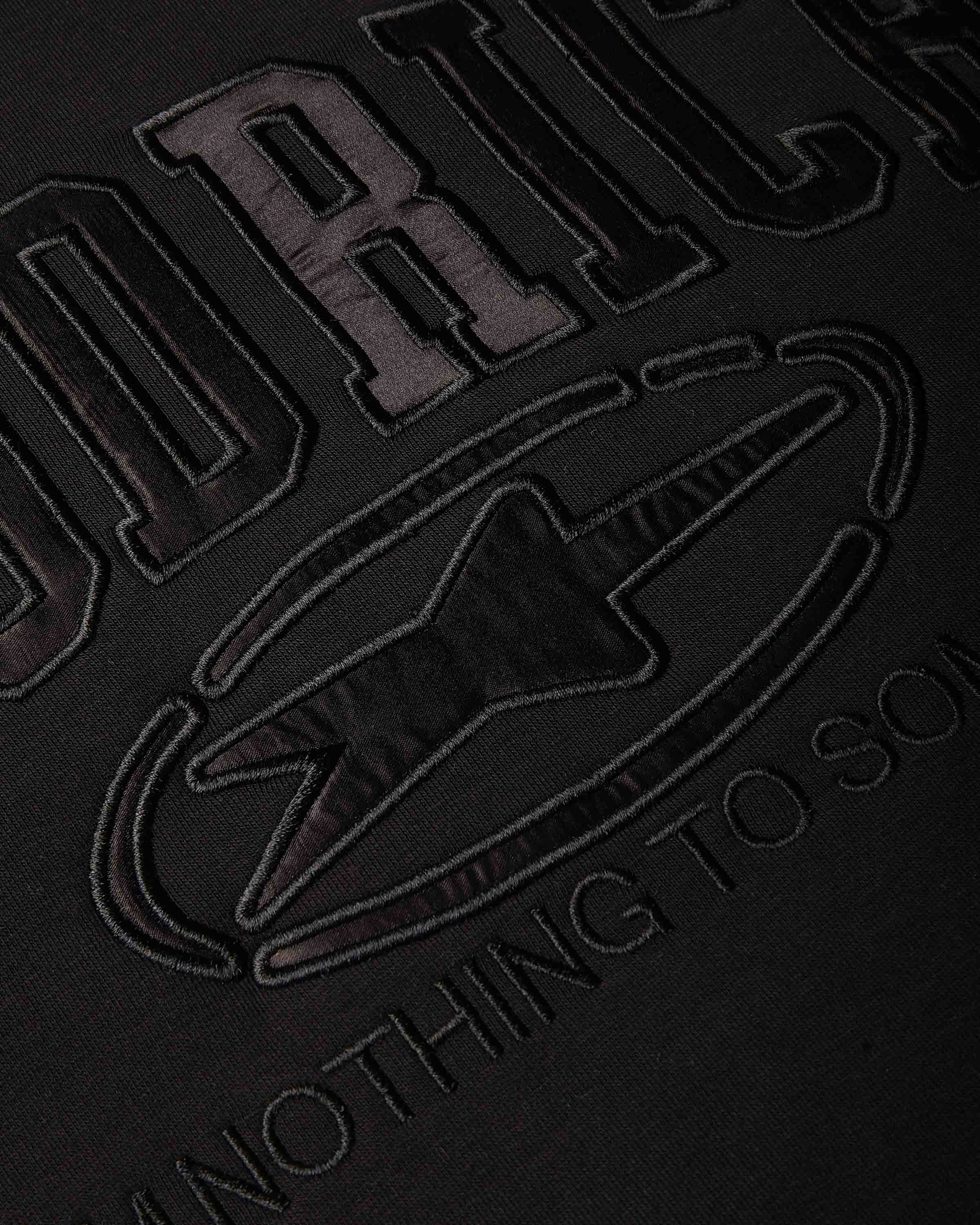 Close-up of a black fabric surface with large tonal embroidered lettering and a star-like emblem, showing partial text.