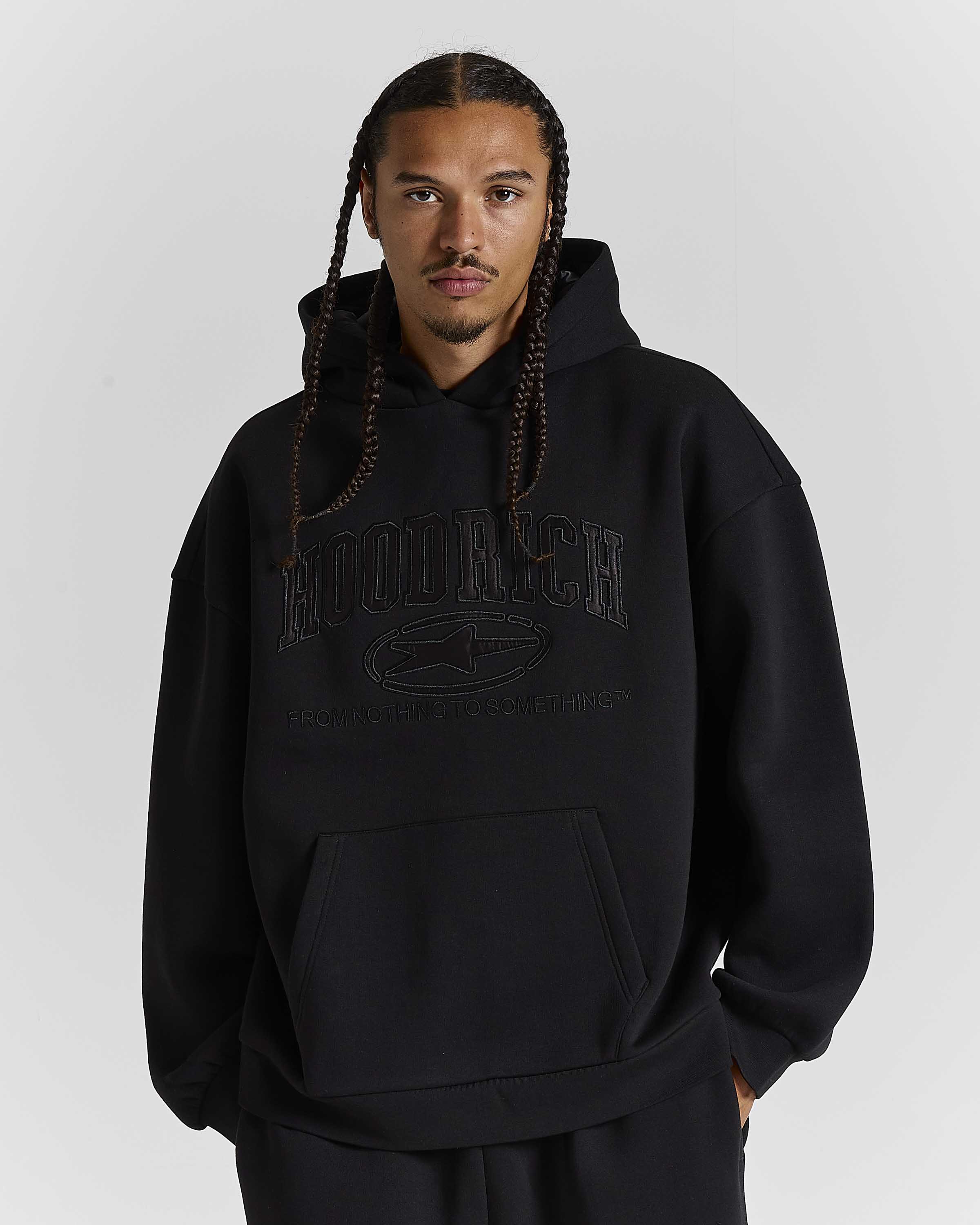 A man with braided hair wearing a black hooded sweatshirt with tonal black graphic text reading 'HOODRICH' and a logo on the chest, photographed against a light gray studio background.