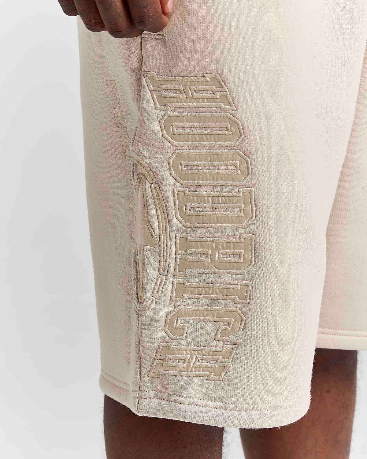 Close-up of a person wearing cream-colored athletic shorts with large tonal embroidered HOODRICH lettering on the side.