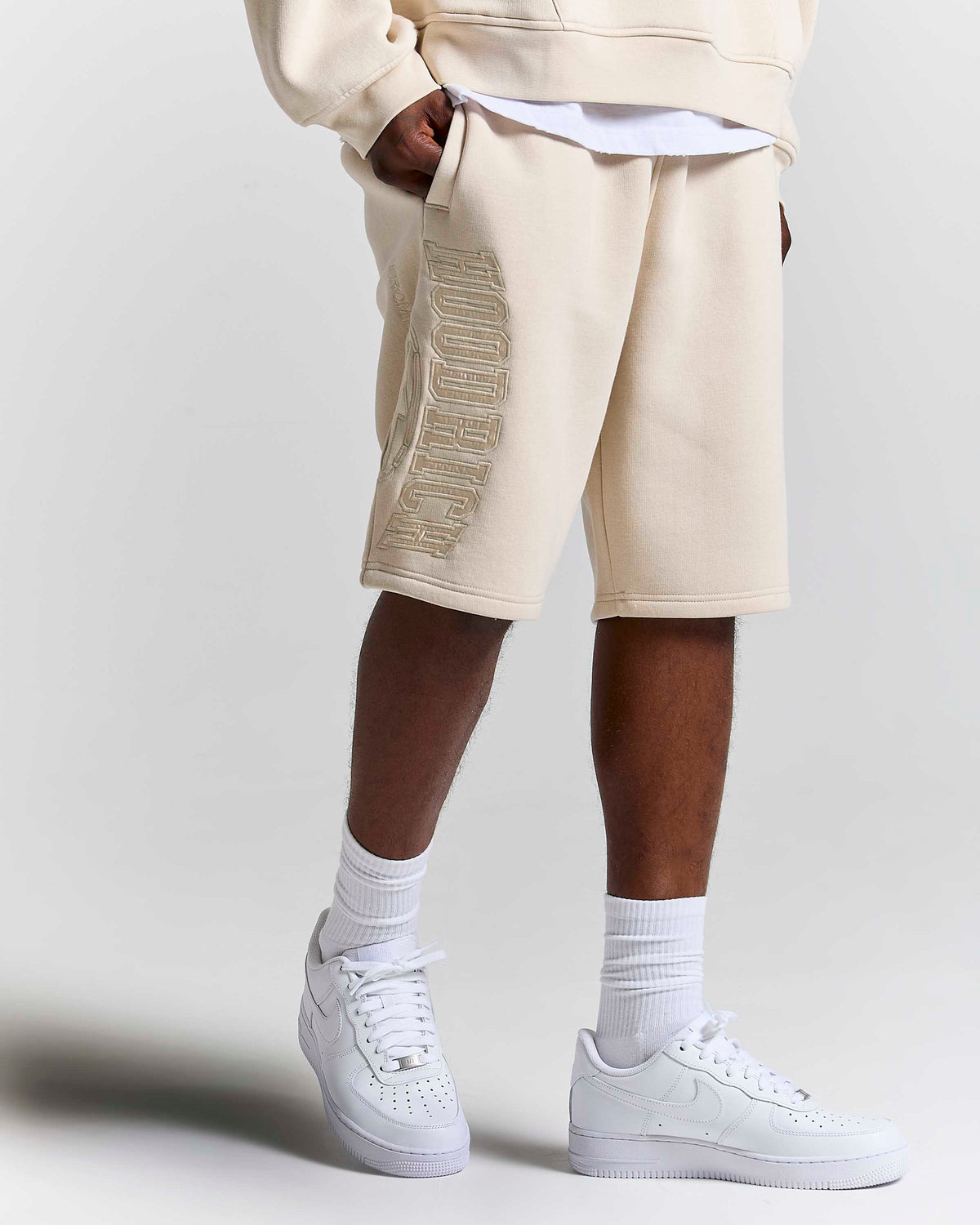 A person wearing beige sweat shorts with a metallic gold embroidered logo down the side, styled with a white t-shirt, white crew socks, and white Nike Air Force 1 sneakers against a light gray studio background.