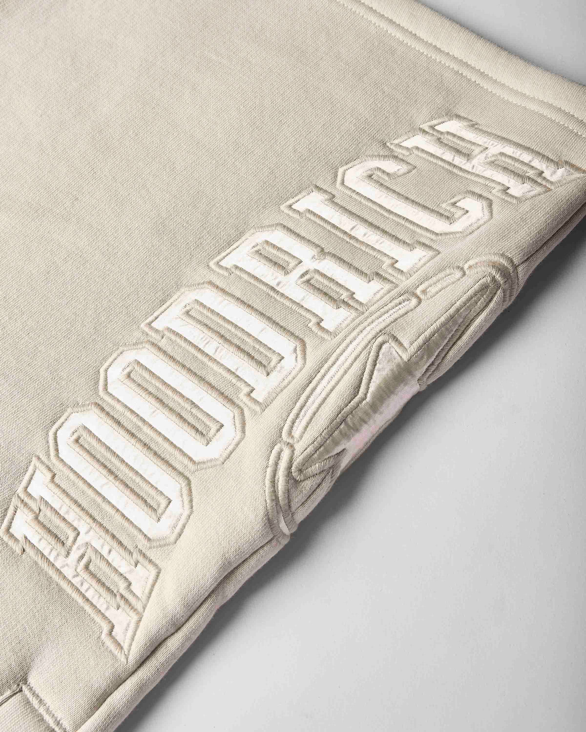 Close-up of a light beige or cream-colored fabric item, likely a sweatshirt or jacket, featuring large raised collegiate-style embroidery reading 'HOODRICH' across the surface.