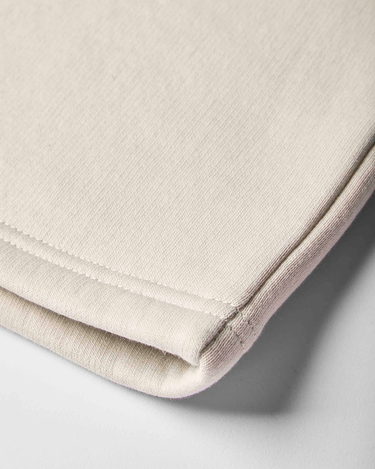 Close-up of a folded light beige fabric item, showing the soft texture and neat stitched edge on a light gray background.