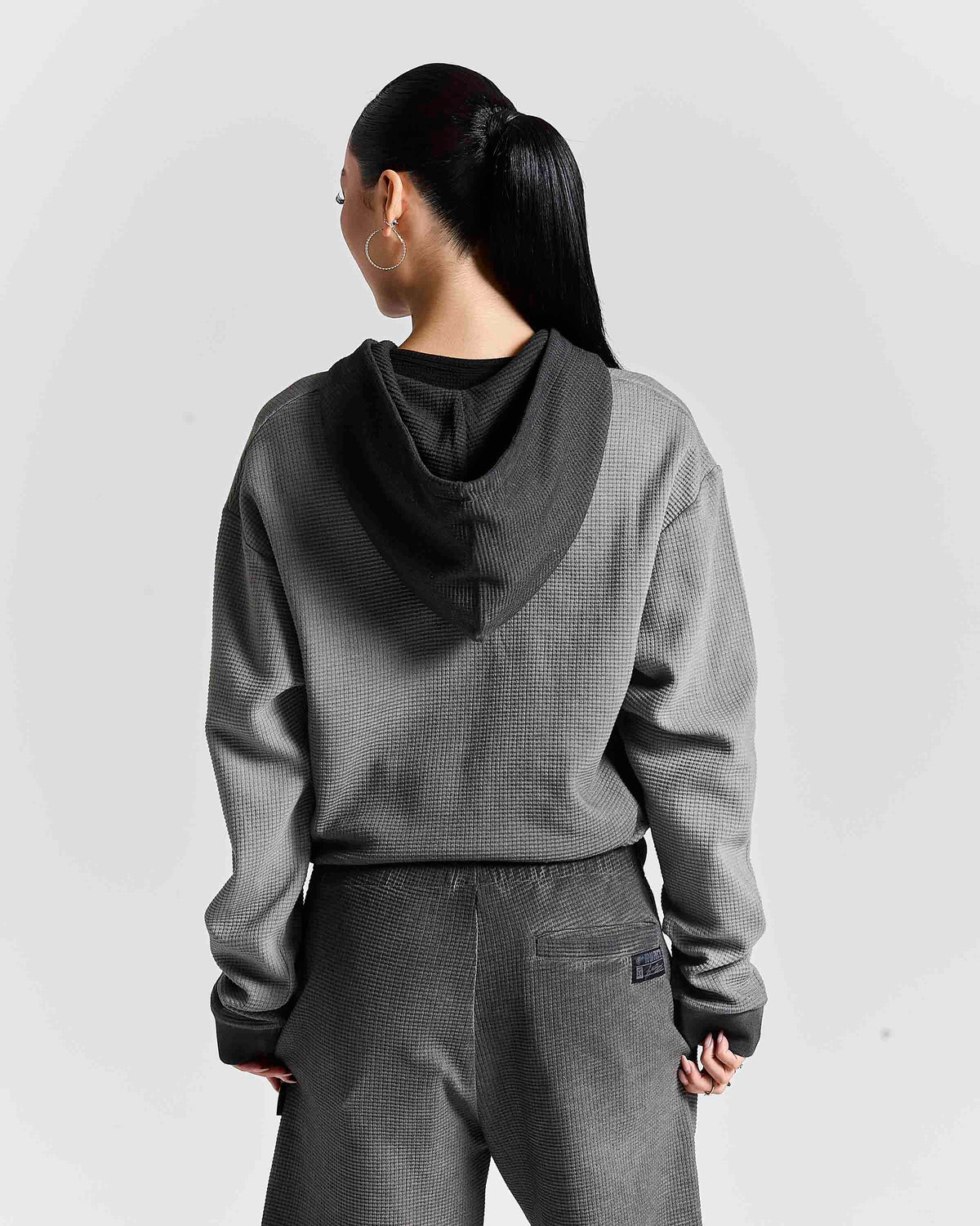 A woman shown from the back wearing a gray textured hooded sweatshirt with a contrasting black inner hood lining and matching gray textured sweatpants, set against a light gray studio background.