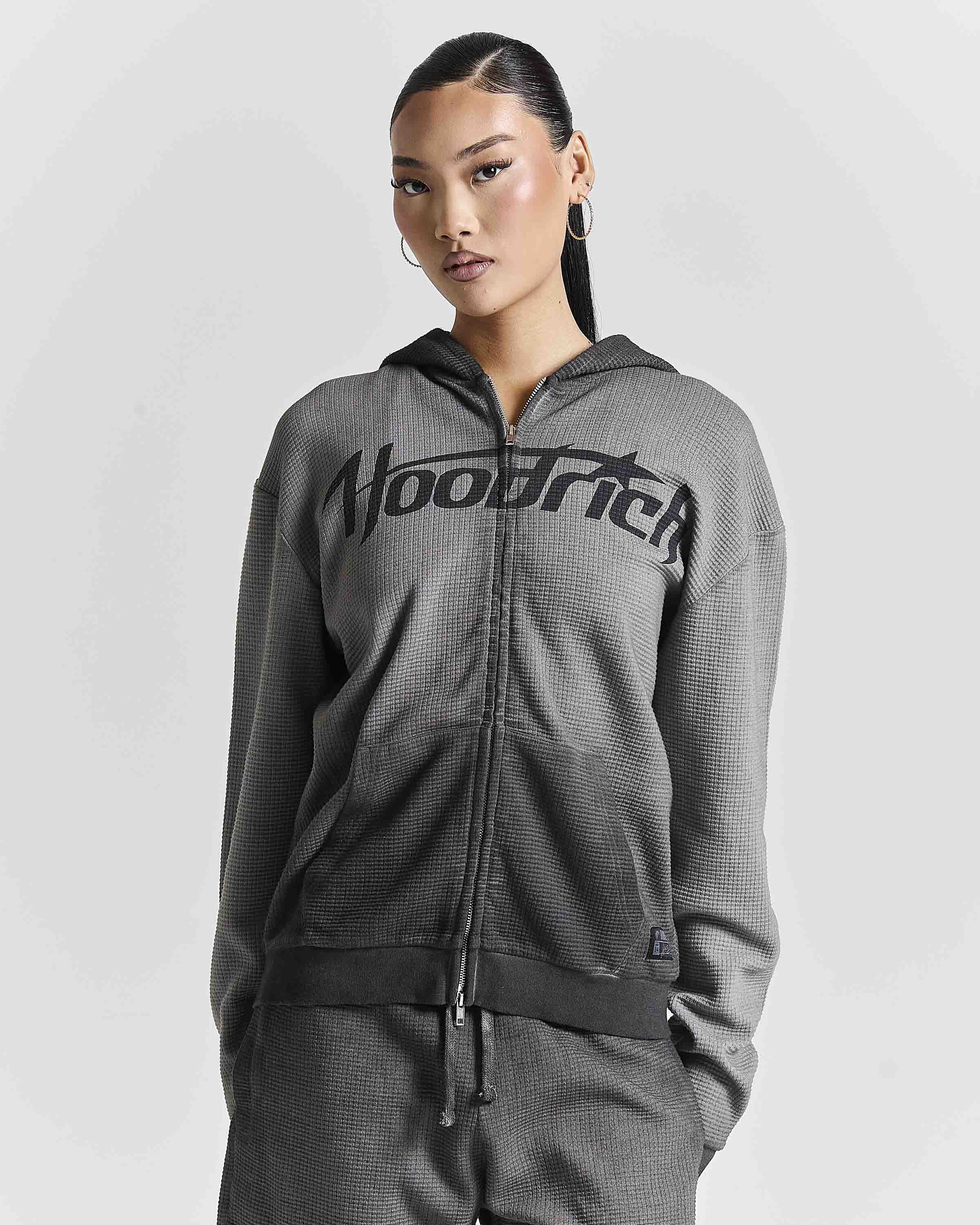 A woman modeling a dark gray waffle-knit zip-up hoodie with a black 'Hoodrich' logo across the chest, paired with matching gray pants, against a light gray studio background.