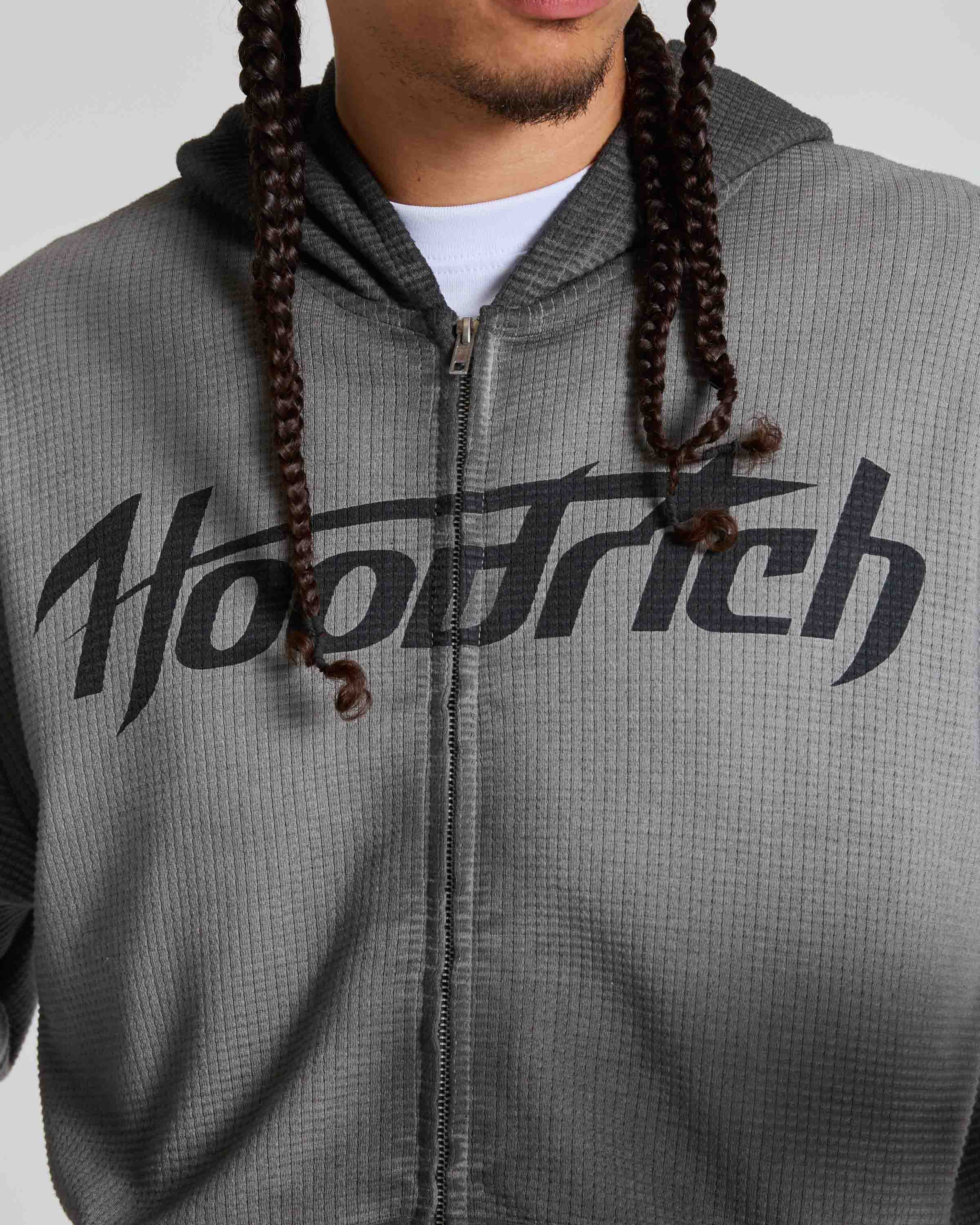 A close-up of a person wearing a gray waffle-knit zip-up hoodie with a large black 'Hoodrich' logo across the chest, over a white t-shirt. The person has dark brown braided hair hanging down the front.
