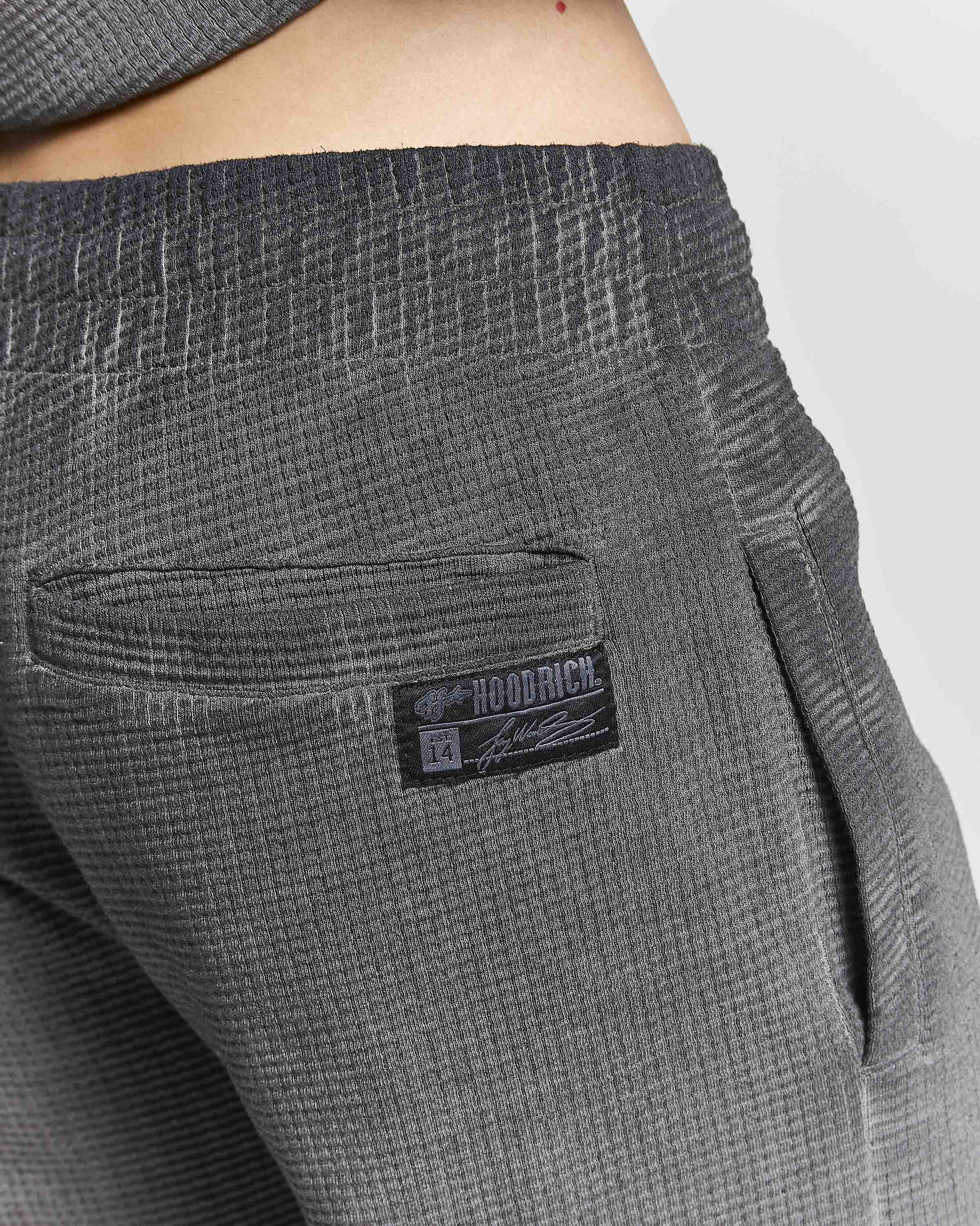 Close-up of dark gray ribbed knit pants showing a side pocket with a black and white HOODRICH brand patch, worn by a model.