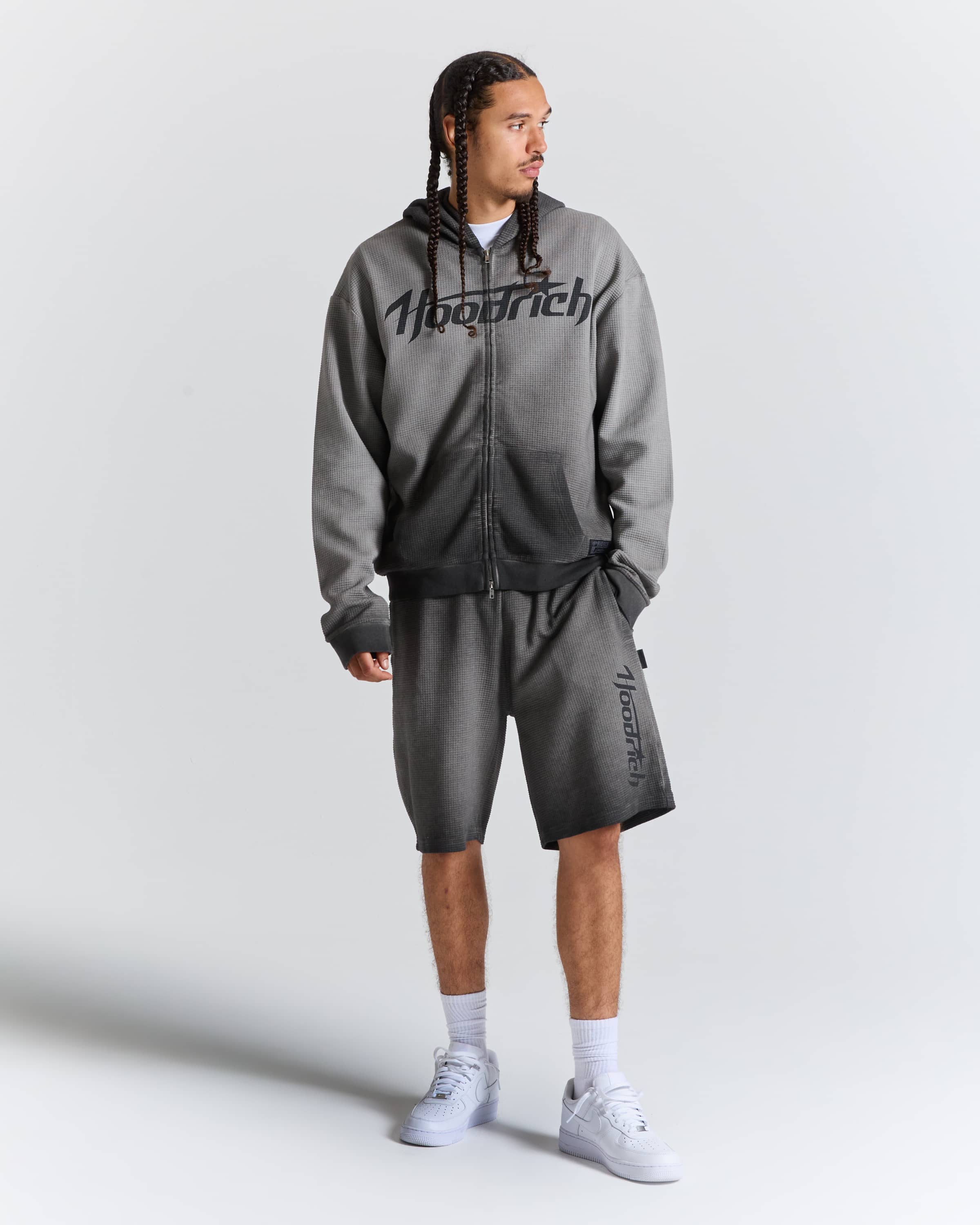 A man modeling a gray zip-up hoodie and matching gray shorts with black 'Hoodrich' branding, styled with white socks and white sneakers against a light gray studio background.