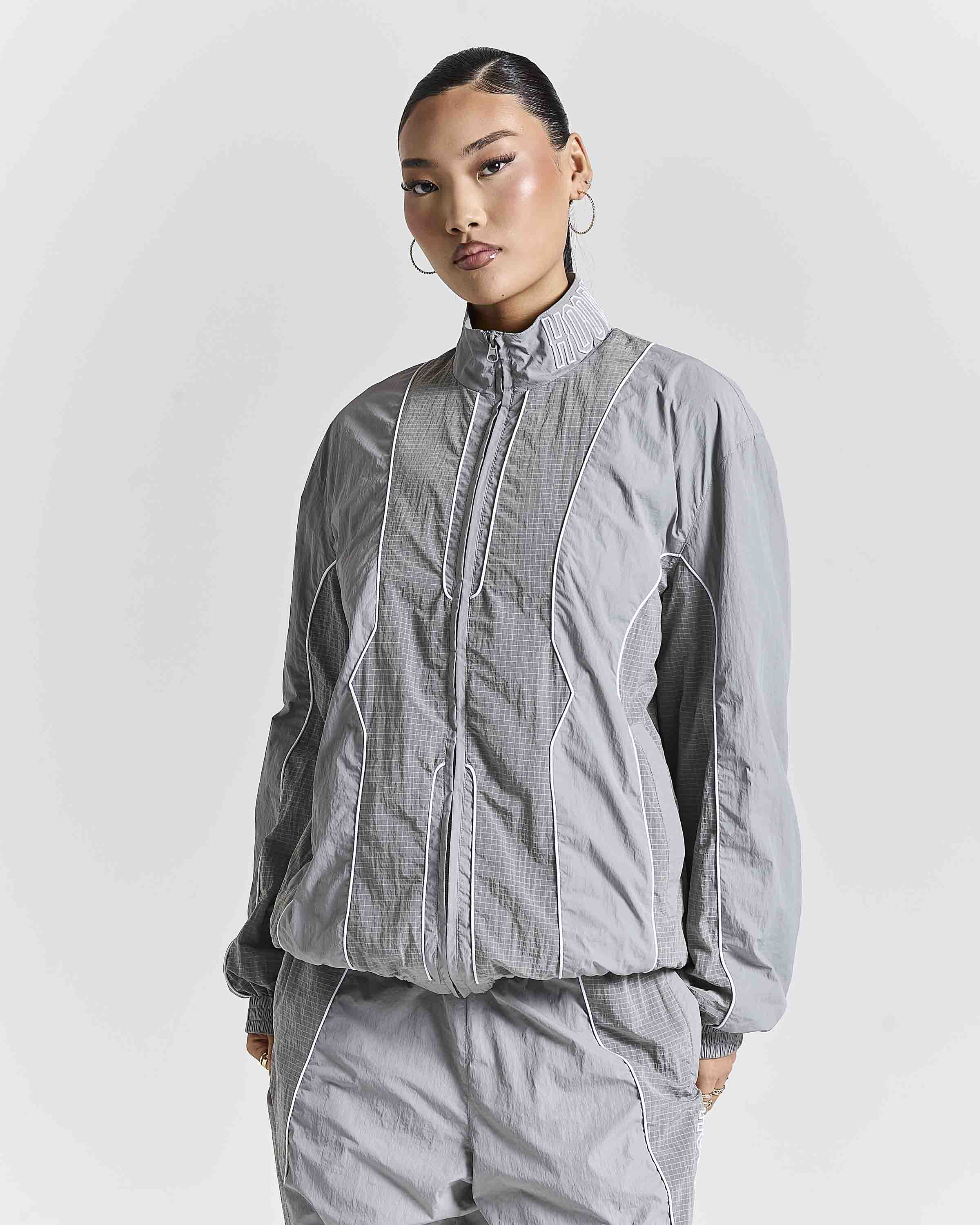 A model wearing a light gray zip-up jacket and matching gray pants against a light gray studio background.