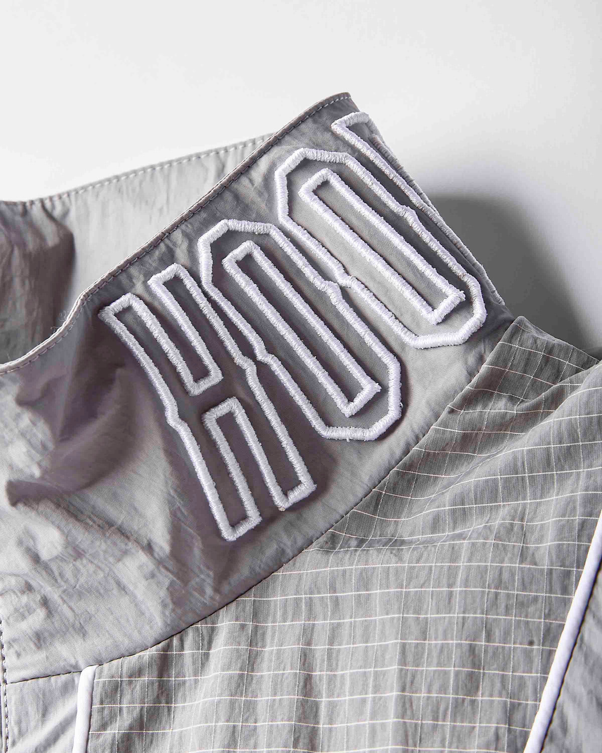 Close-up of a gray lightweight fabric garment with a high collar featuring large white embroidered lettering outlined in black.