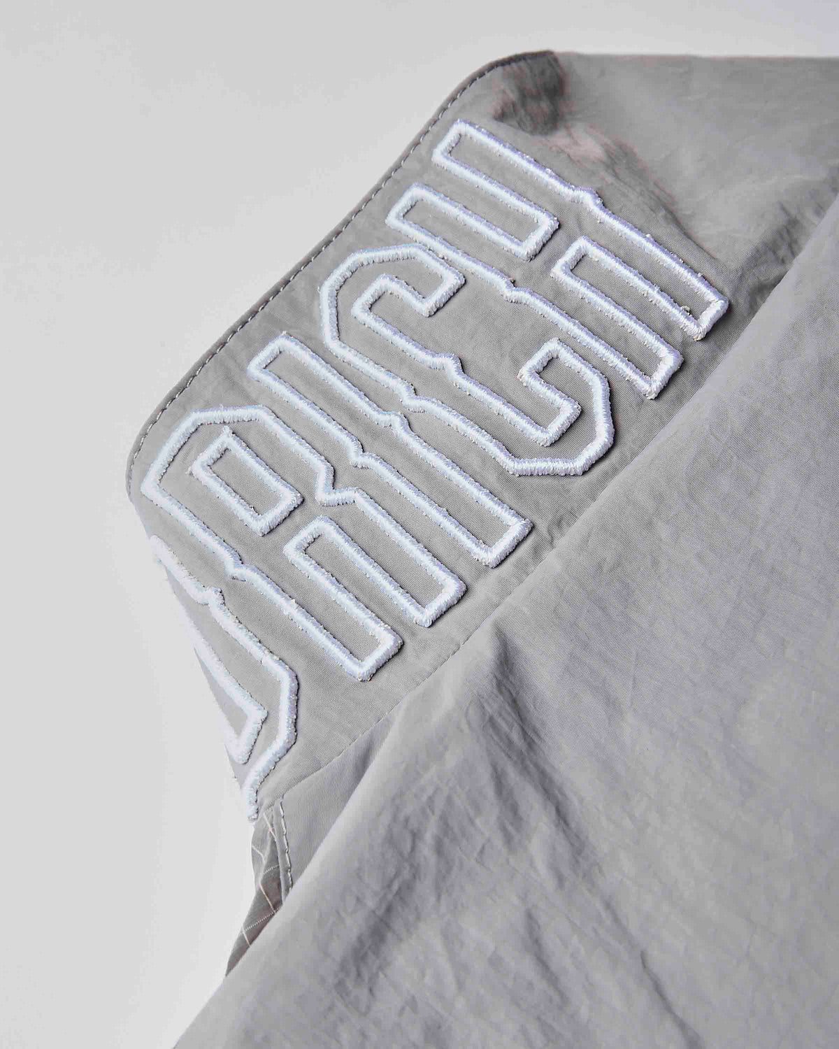 Close-up of a light gray fabric garment with large white embroidered lettering reading 'RICH' on the shoulder.