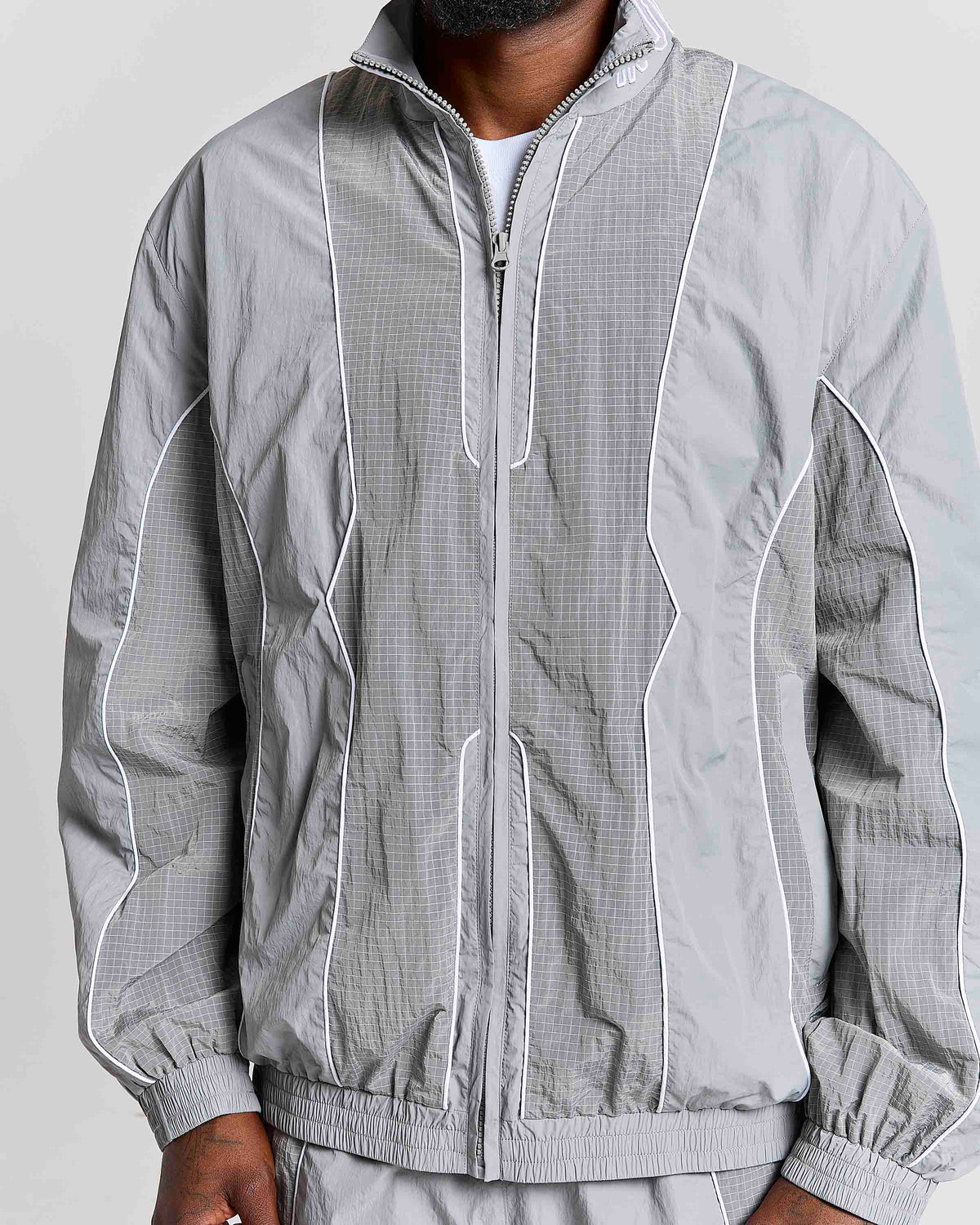 A close-up of a person wearing a light gray zip-up windbreaker jacket with white piping and a subtle grid texture, paired with matching gray pants against a plain light background.