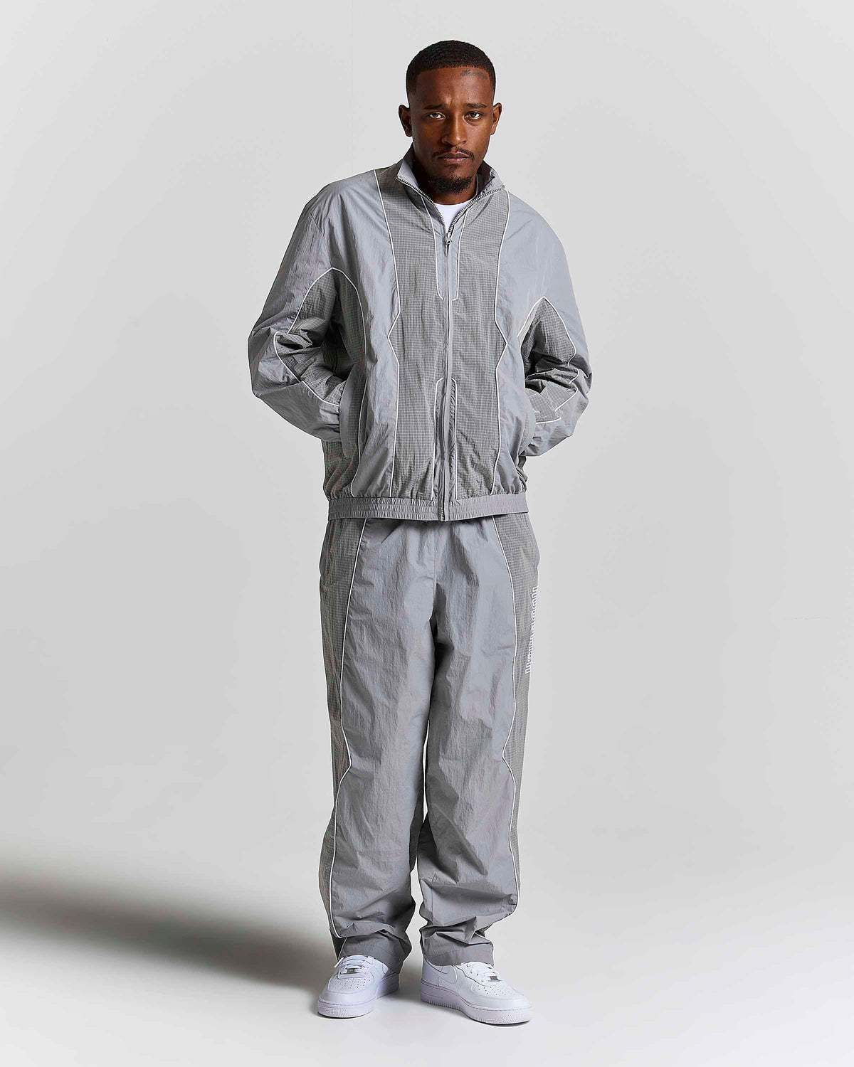 A man modeling a light gray two-piece tracksuit consisting of a lightweight zip-up jacket and matching jogger pants, paired with white sneakers, against a plain light gray studio background.