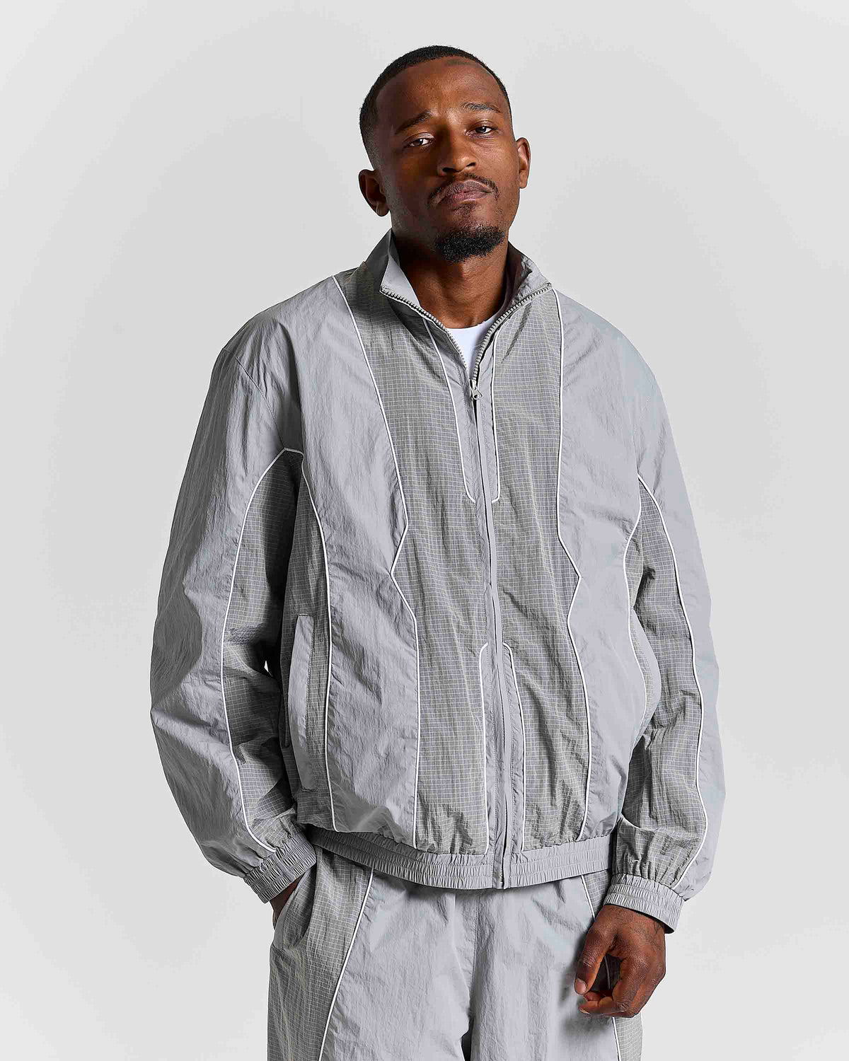 A man wearing a light gray zip-up windbreaker jacket with matching light gray trousers, photographed against a plain light gray studio background.