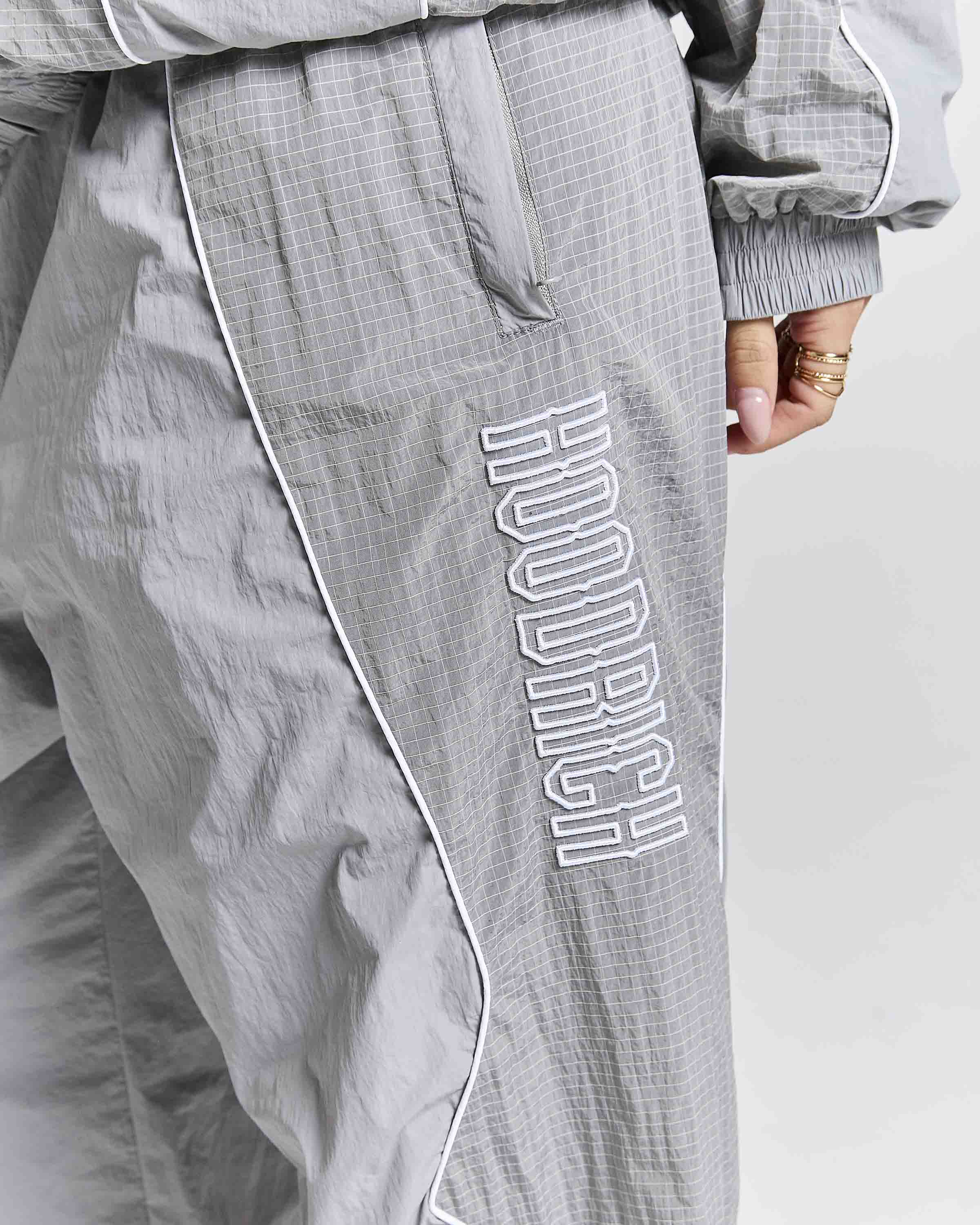 Close-up of gray textured windbreaker pants featuring a large white outlined 'HOODRICH' logo printed vertically on the left leg, with white piping and a visible hand wearing rings.