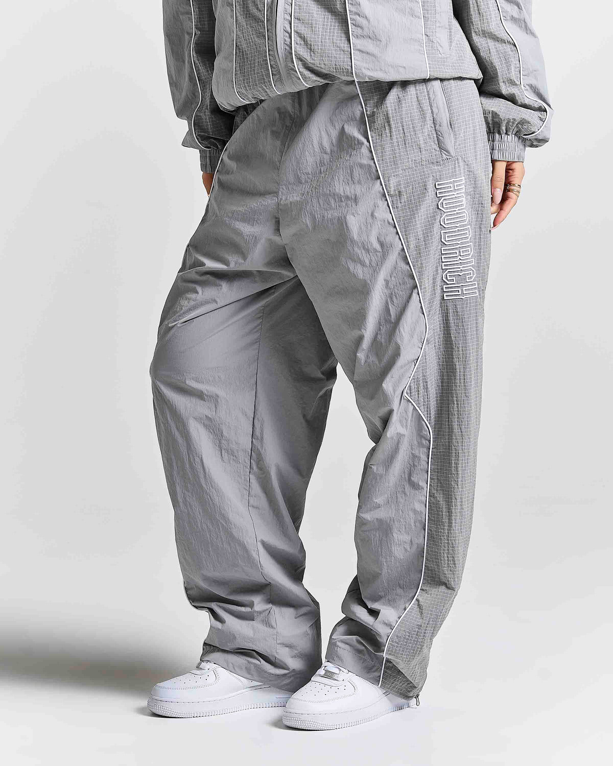 A person wearing loose-fit gray pants with a subtle grid texture, white sneakers, and a matching gray jacket against a light gray studio background.