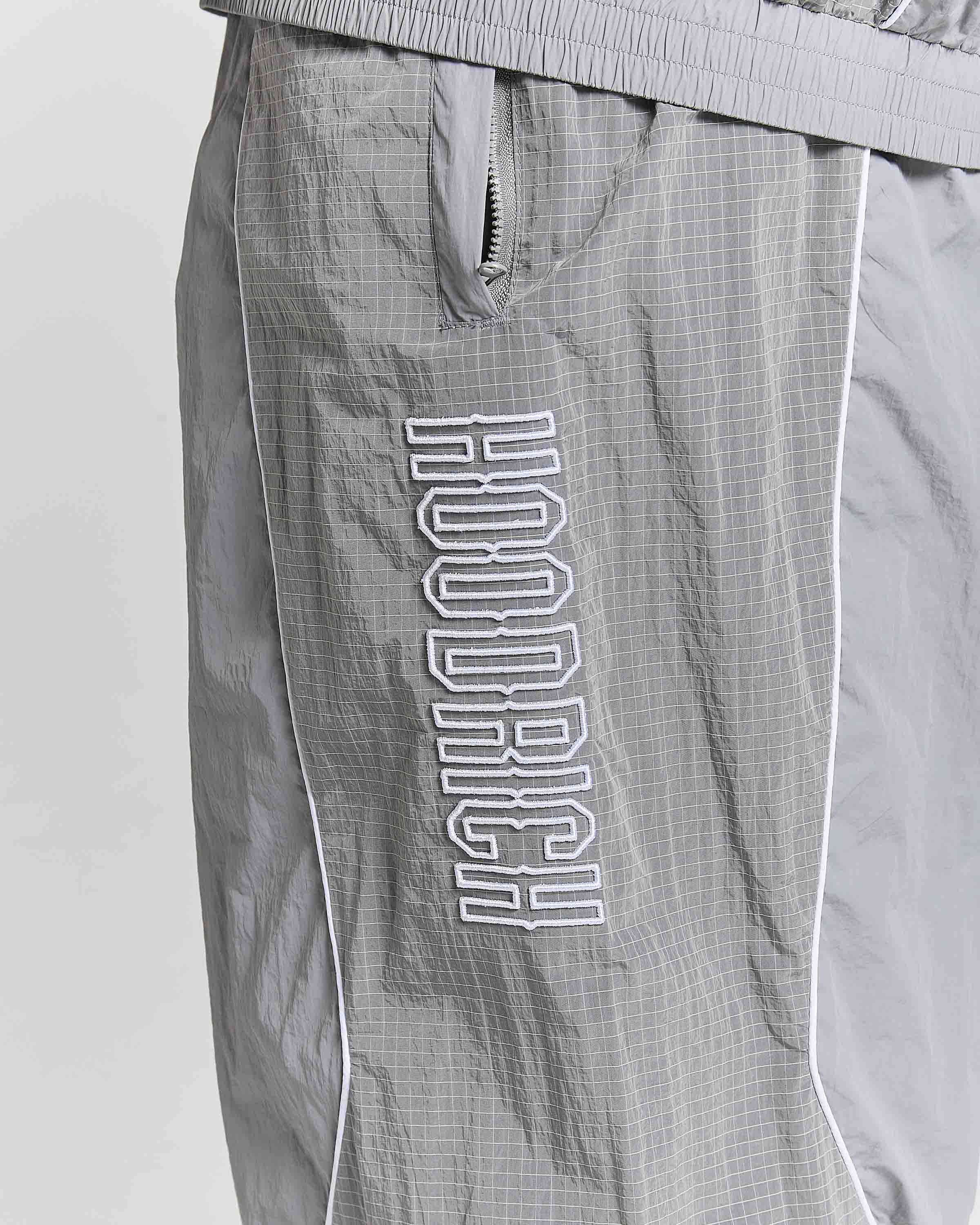 Close-up of gray HOODRICH windbreaker shorts with a white outlined vertical logo on the leg, featuring a zippered pocket and white piping.