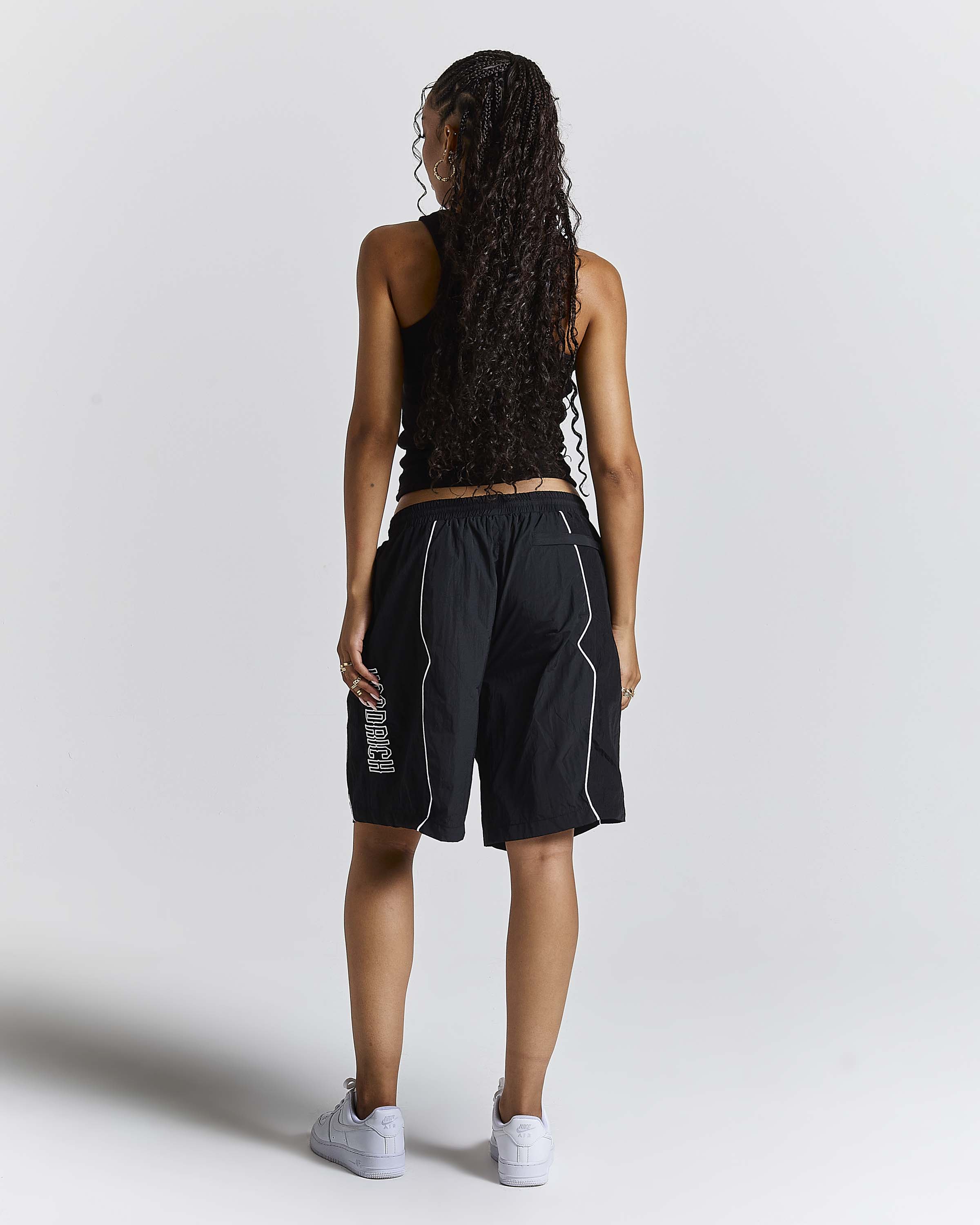 A woman is shown from behind wearing black athletic shorts with white piping and a white vertical logo on the left leg, paired with a black tank top and white sneakers, against a light gray studio background.
