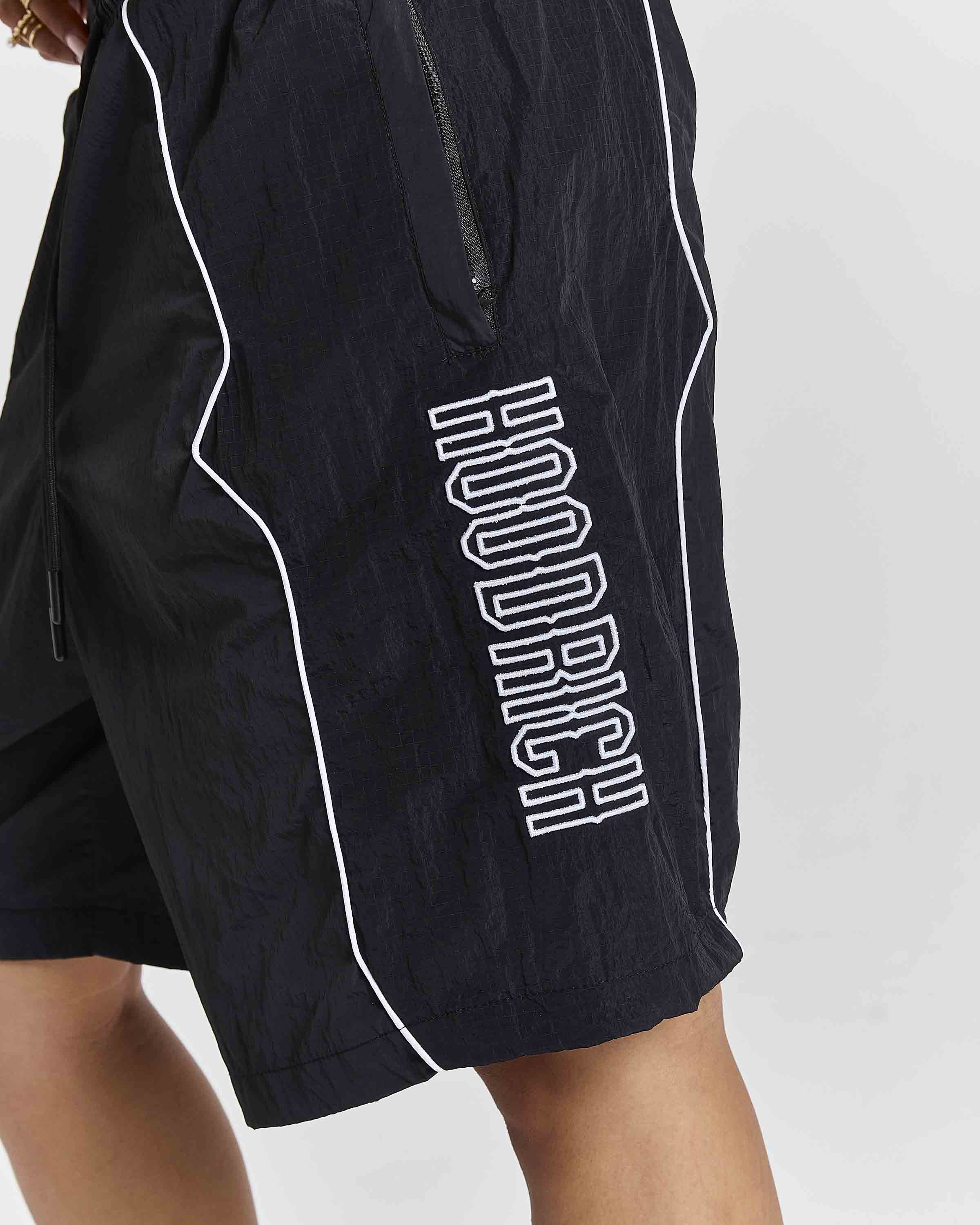 Close-up of black athletic shorts with white piping and a large white outlined vertical 'HOODRICH' logo on the side, worn by a model against a light gray background.