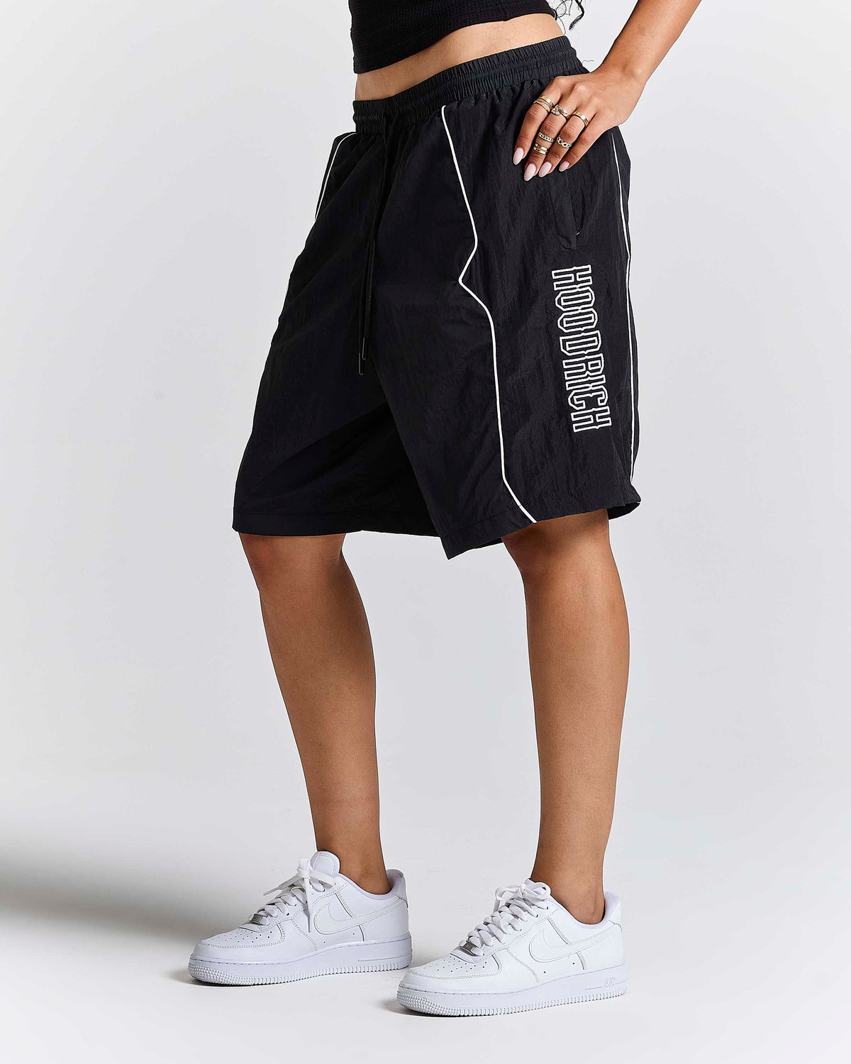 A person wearing black athletic shorts with white piping and vertical white 'HOODRICH' text on the side, paired with white sneakers, photographed against a light gray studio background.
