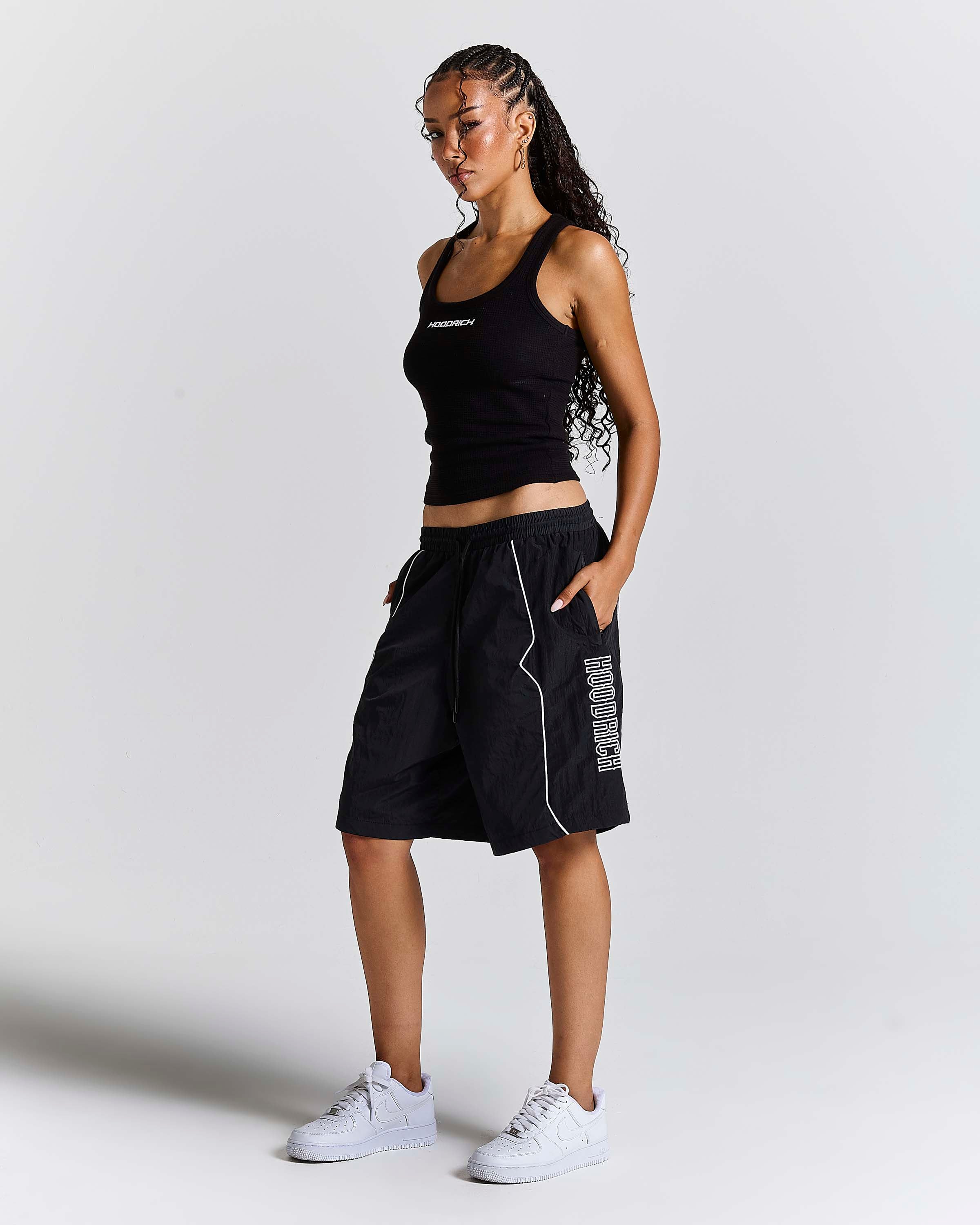 A woman modeling black athletic wear, featuring a black ribbed tank top and black shorts with white piping and white side text, paired with white sneakers, against a light gray studio background.