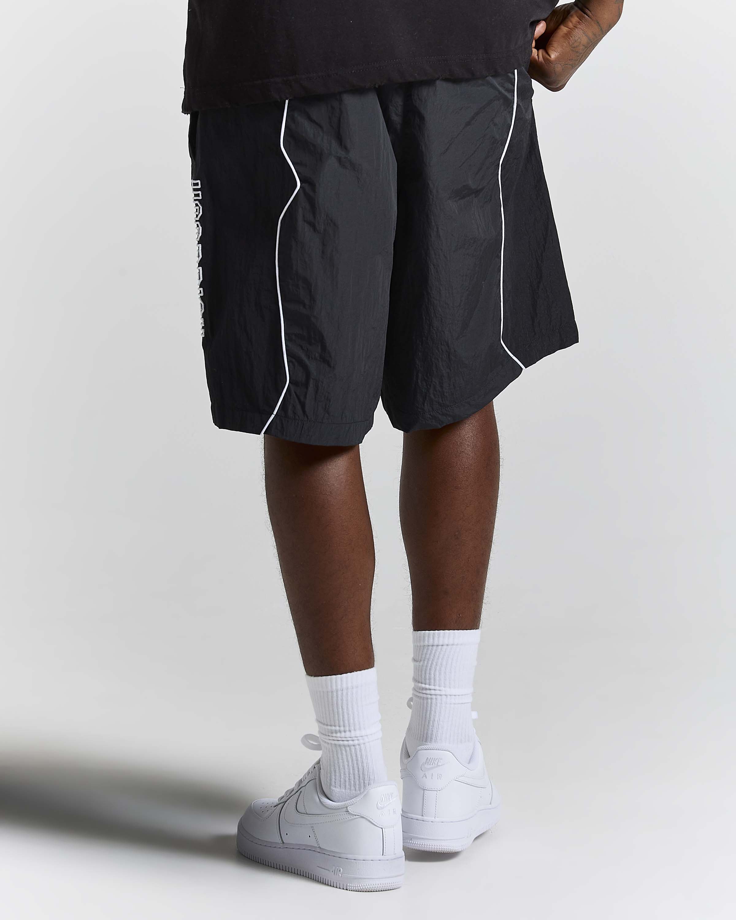 A person shown from the waist down wearing black athletic shorts with white piping, white crew socks, and white sneakers against a light gray studio background.