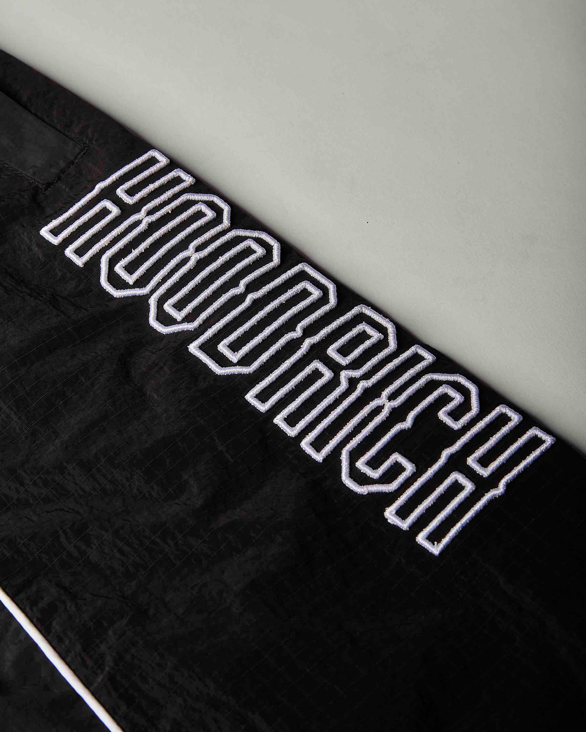 Close-up of a black textured fabric item featuring a large white embroidered 'HOODRICH' logo in a varsity-style outline font.