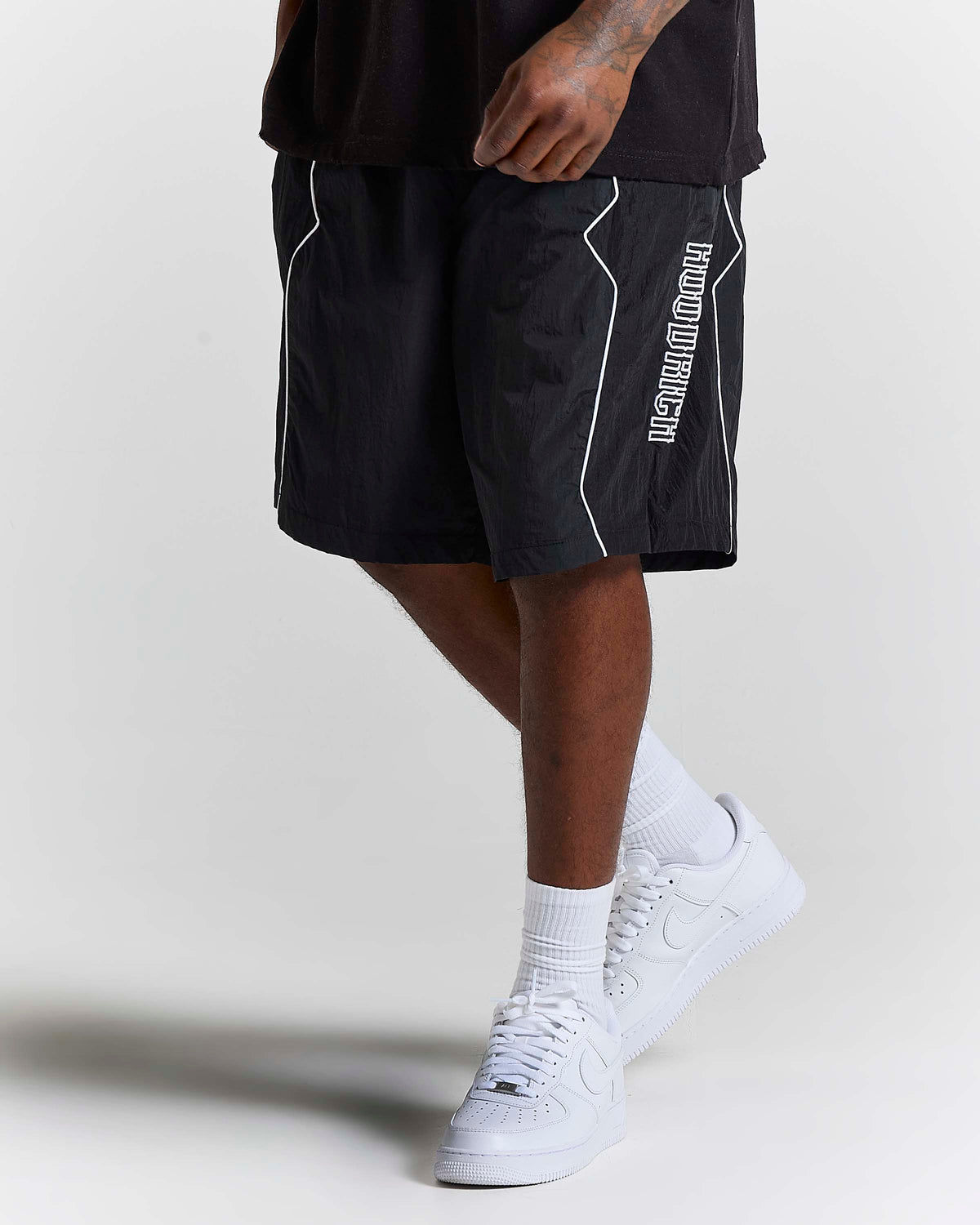 A person wearing black athletic shorts with white piping and vertical white 'HOODRICH' text on the side, paired with white socks and white sneakers, photographed against a light gray studio background.