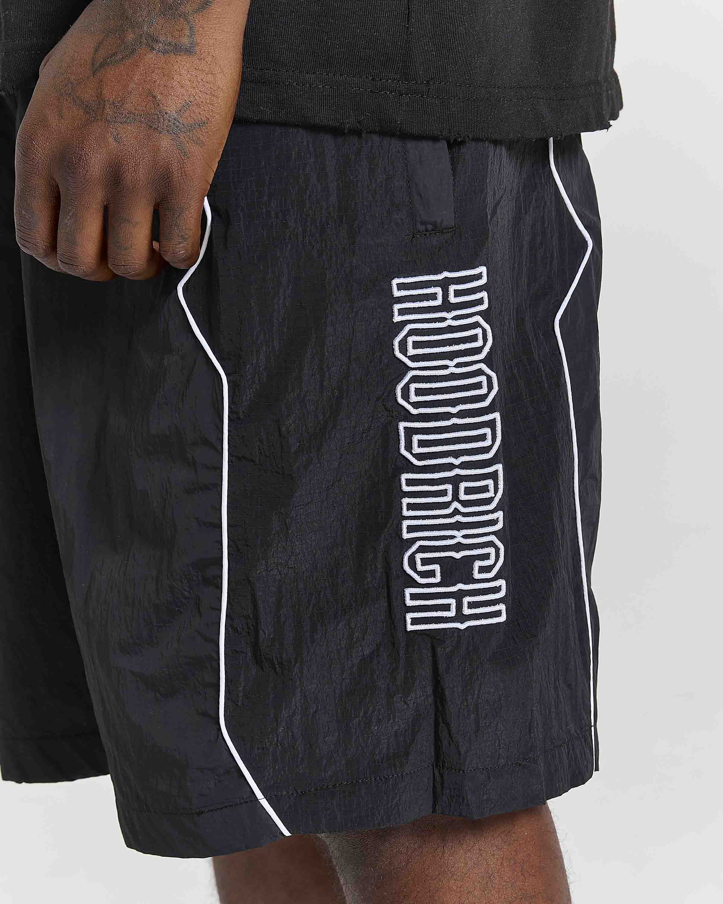 Close-up of a person wearing black athletic shorts with white piping and a large vertical white 'HOODRICH' logo on the side, paired with a black shirt against a light gray background.