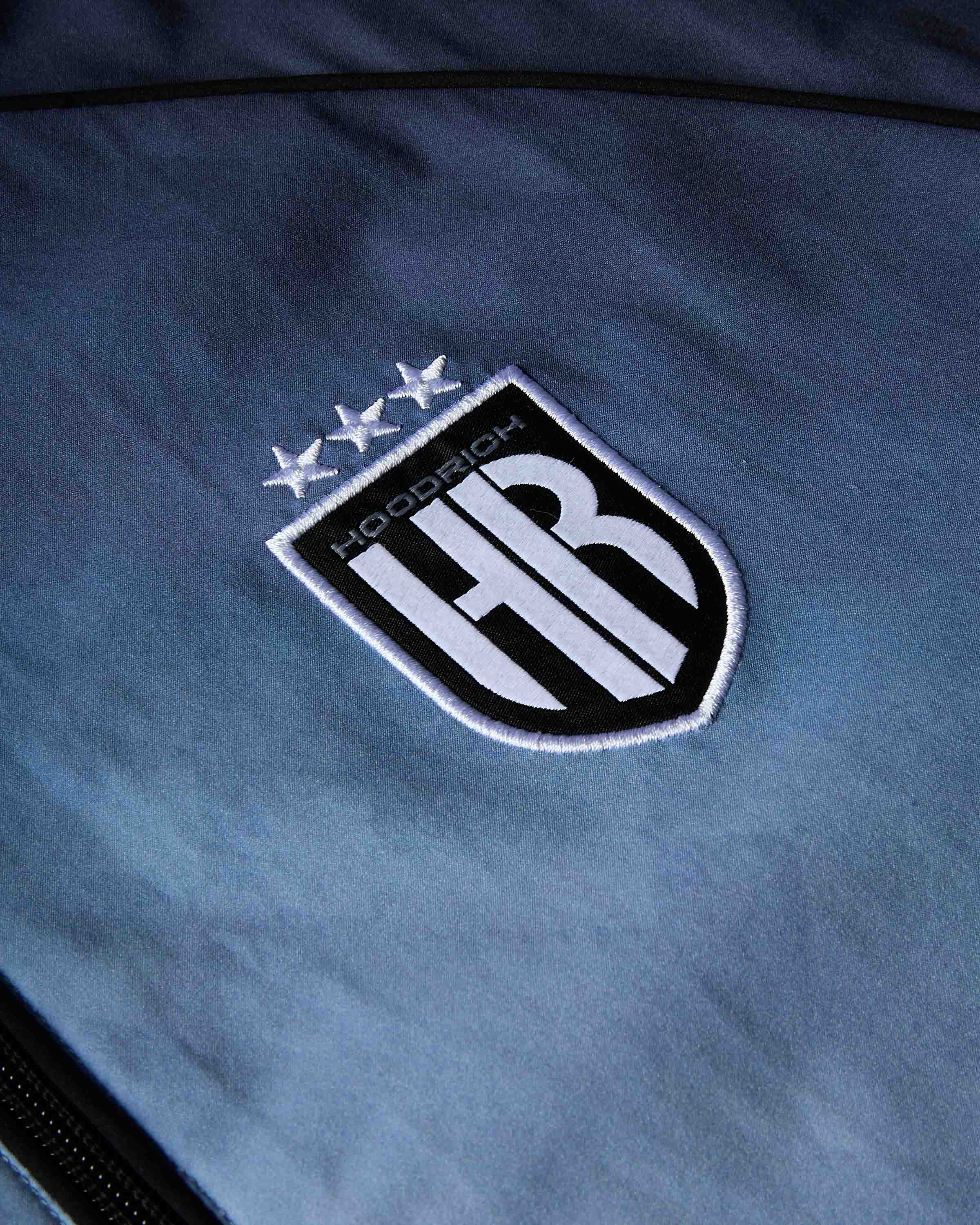 Close-up of a blue fabric surface featuring a black and white embroidered shield crest with three stars and the word 'HOODRICH' above large stylized letters.