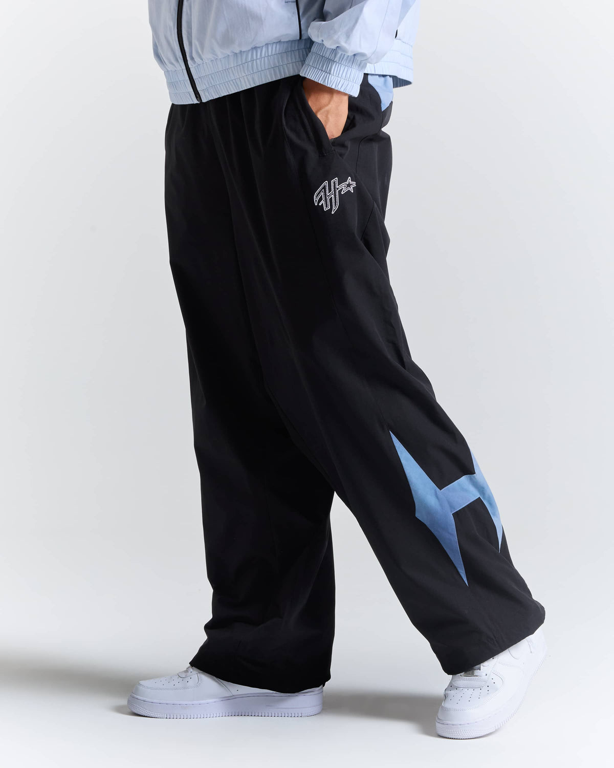 A person wearing black wide-leg sweatpants with a light blue graphic on the lower leg, paired with a light blue zip jacket and white sneakers, shown from the waist down against a light gray studio background.