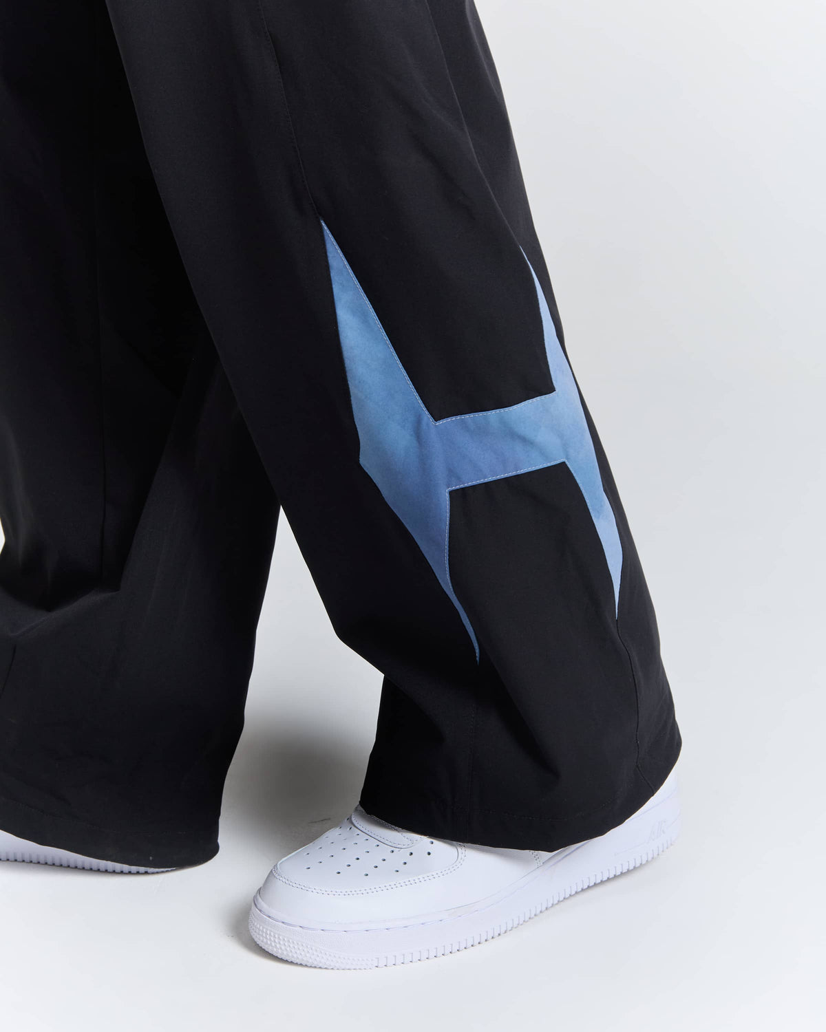 Close-up of a person's lower legs wearing black wide-leg pants with a large light blue geometric star graphic on the outer side and white low-top sneakers, against a light gray studio background.