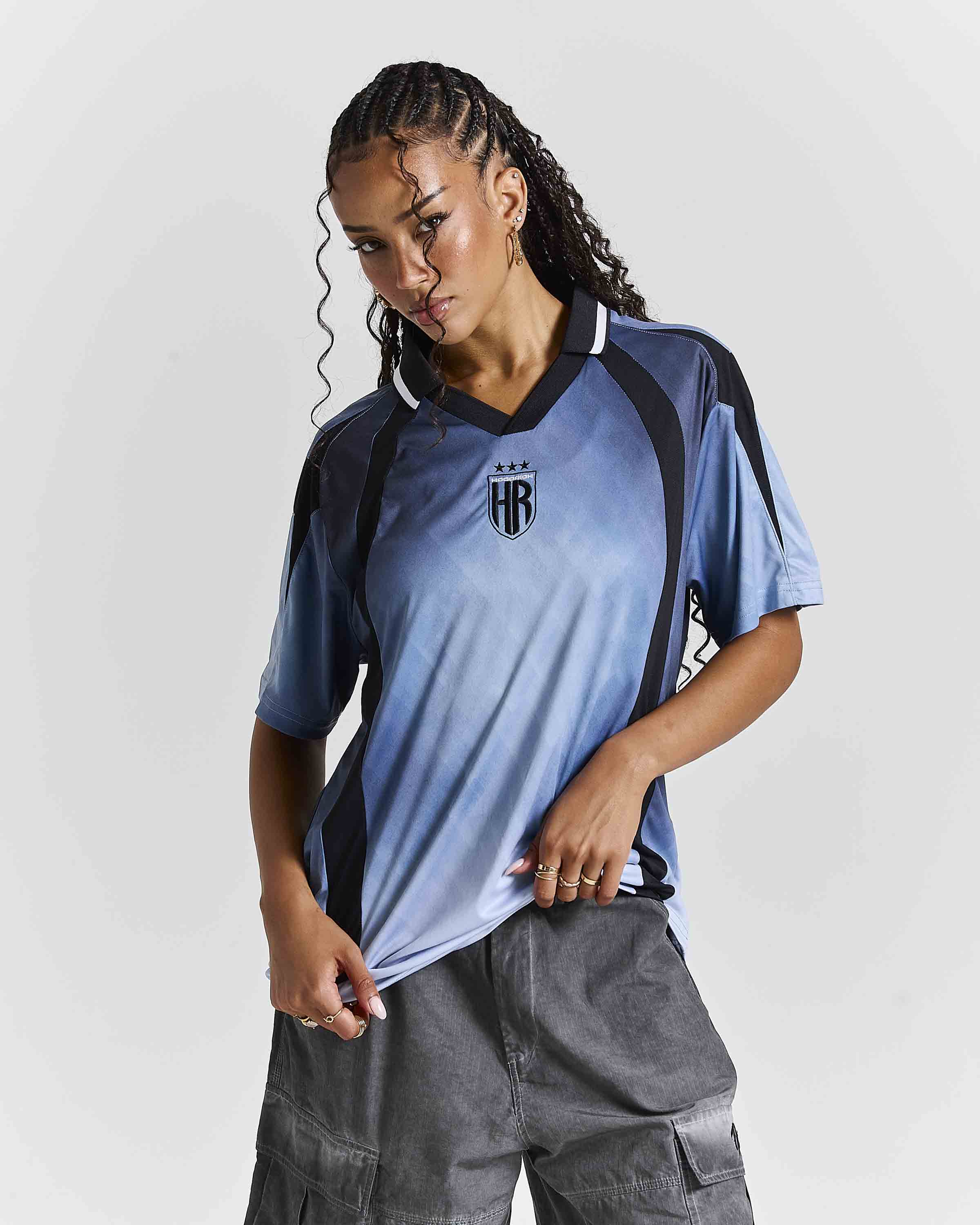 A woman modeling a short-sleeve athletic jersey with a blue-to-light-blue gradient, black side panels, and a black crest on the chest, paired with gray cargo pants against a light gray studio background.