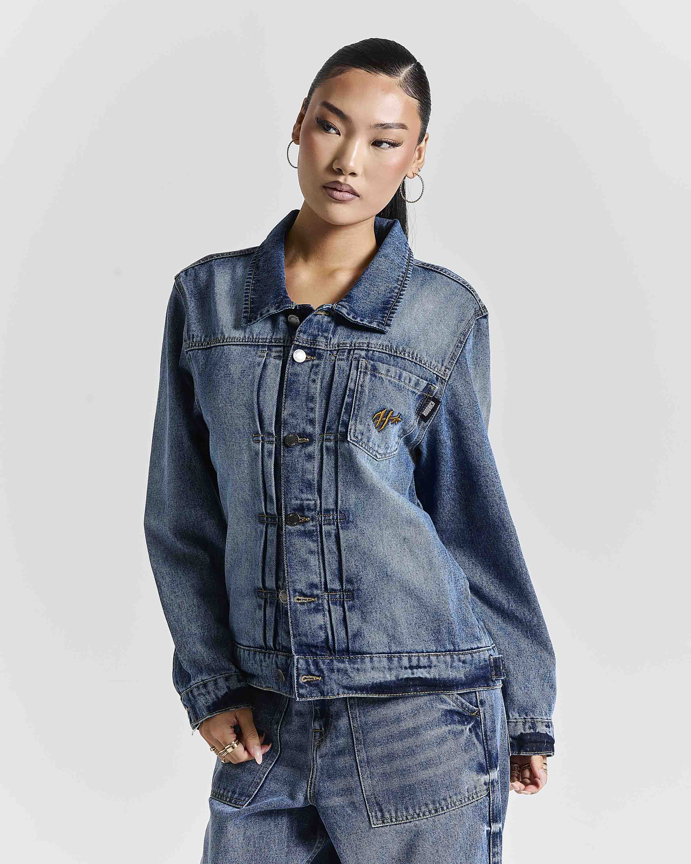 A model wearing a blue denim jacket with a collar, button front, and a small embroidered logo on the chest pocket, paired with matching blue denim pants against a light gray studio background.