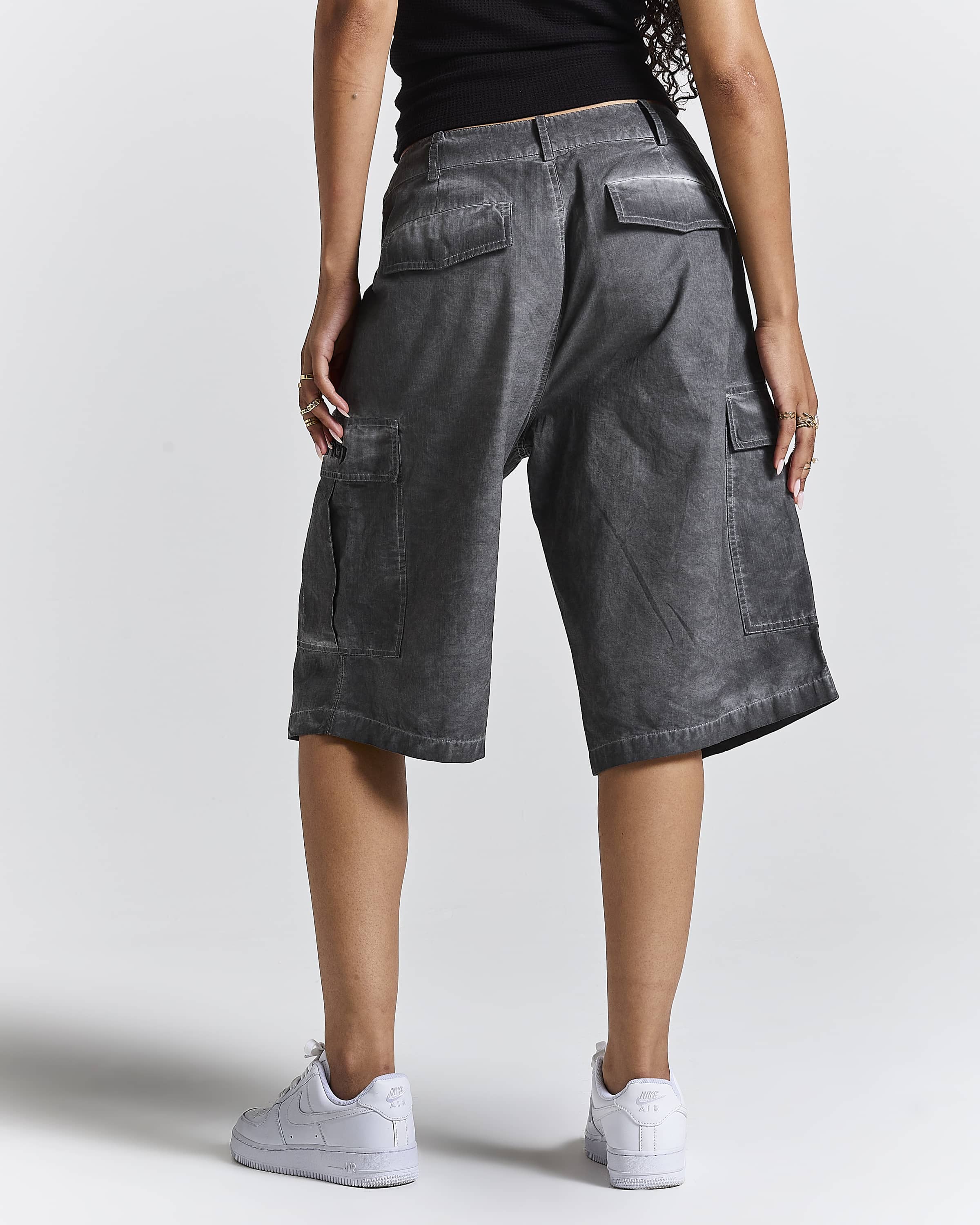 A person shown from the back wearing washed charcoal gray cargo shorts, a black top, and white sneakers against a light gray studio background.