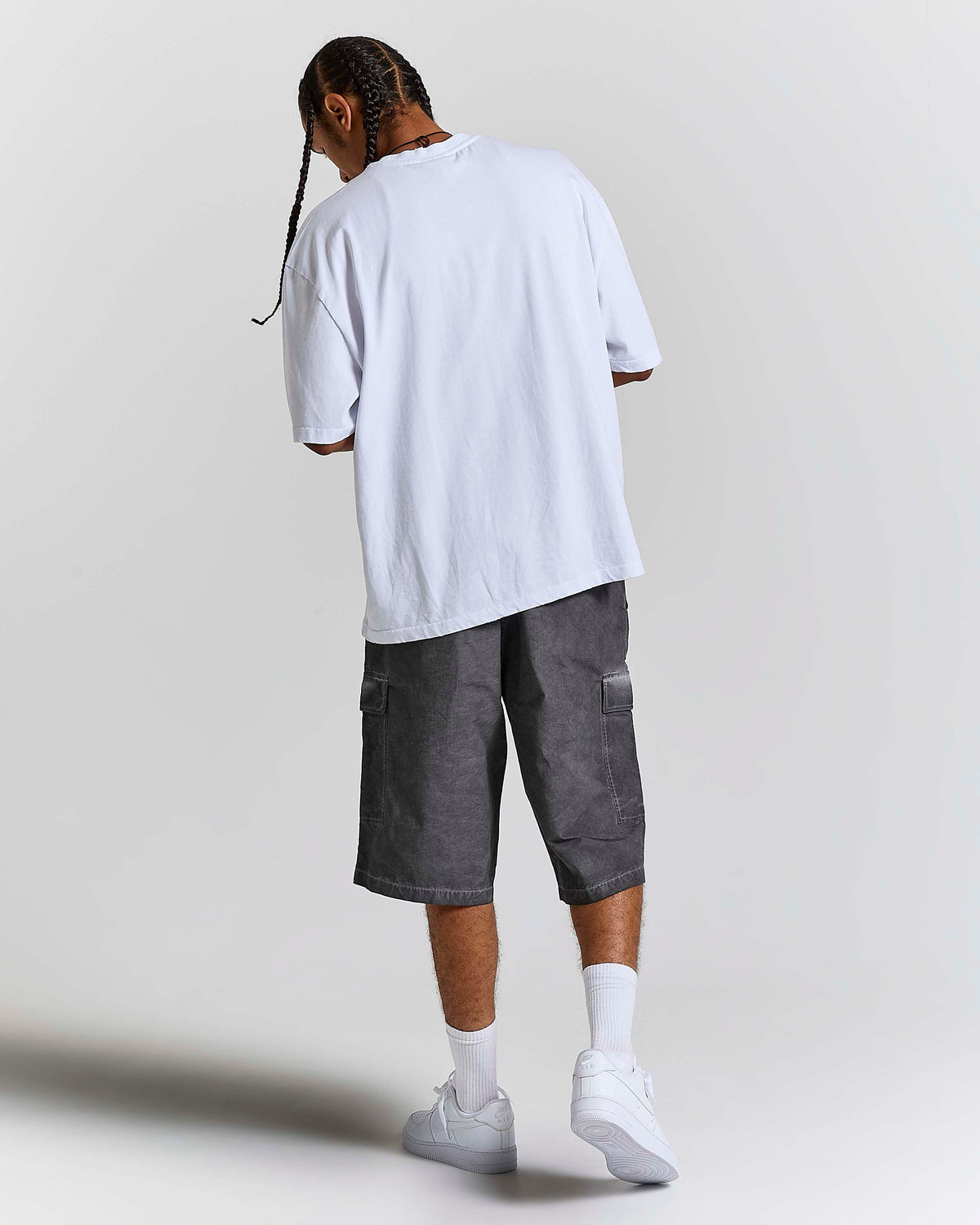 A man shown from behind wearing an oversized white t-shirt, dark gray cargo shorts, white socks, and white sneakers, standing against a light gray studio background.