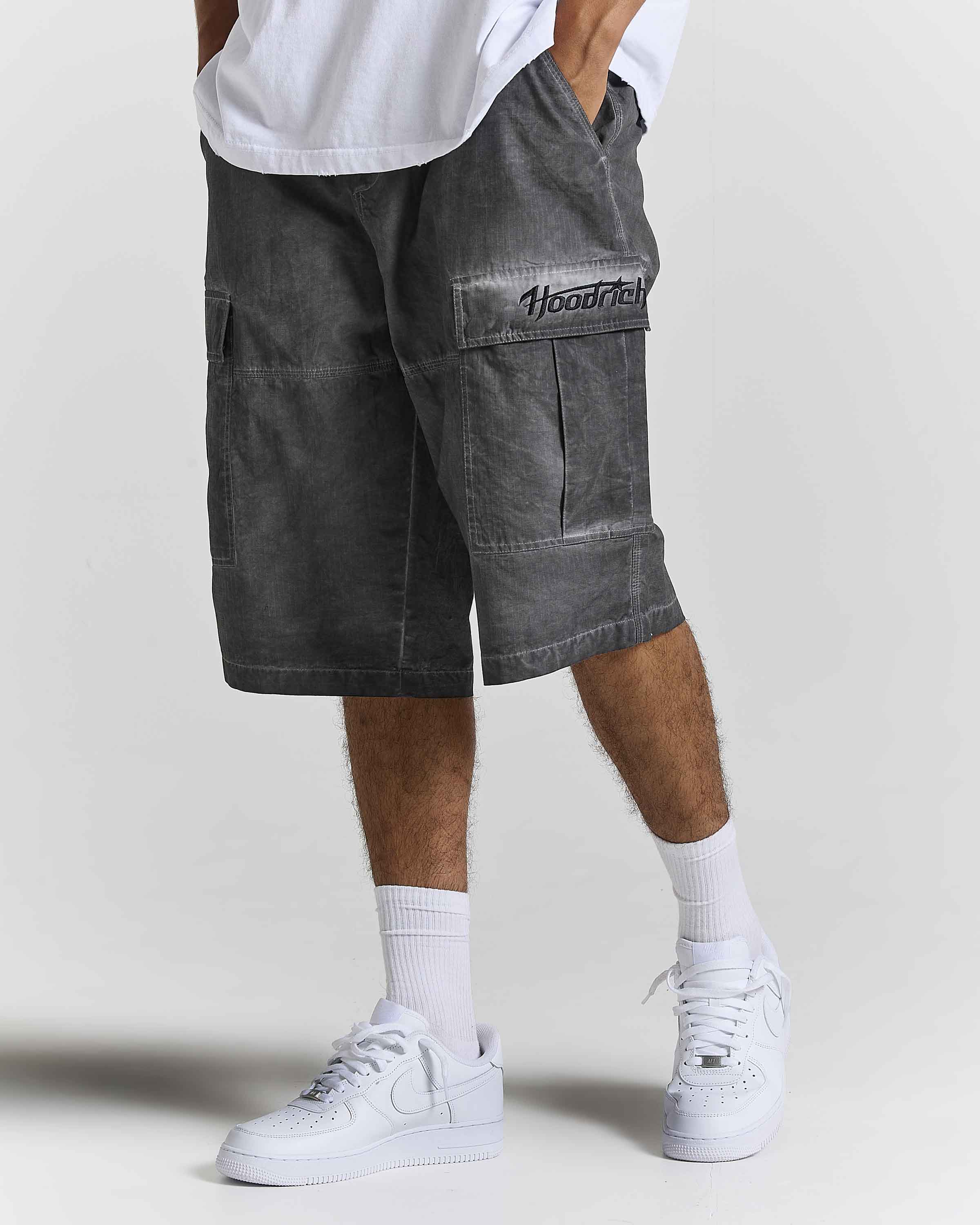 A person wearing dark gray washed cargo shorts with a black embroidered 'Hoodrich' logo on the pocket, styled with a white t-shirt, white socks, and white sneakers against a light gray studio background.