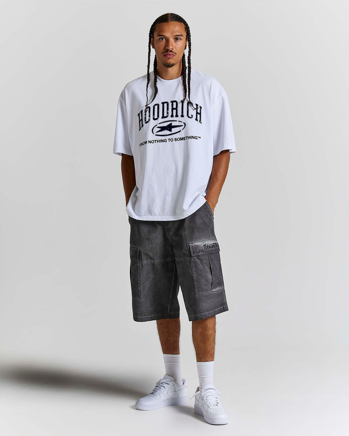 A man modeling an oversized white Woodrich t-shirt with a black graphic print, paired with dark gray cargo shorts, white socks, and white sneakers, standing against a light gray studio background.