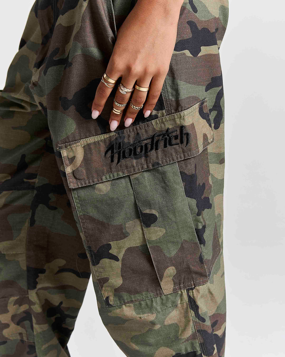 Close-up of a person wearing camouflage cargo pants, with a hand resting on the thigh over a green flap pocket that has the word 'Hoodrich' embroidered in black.