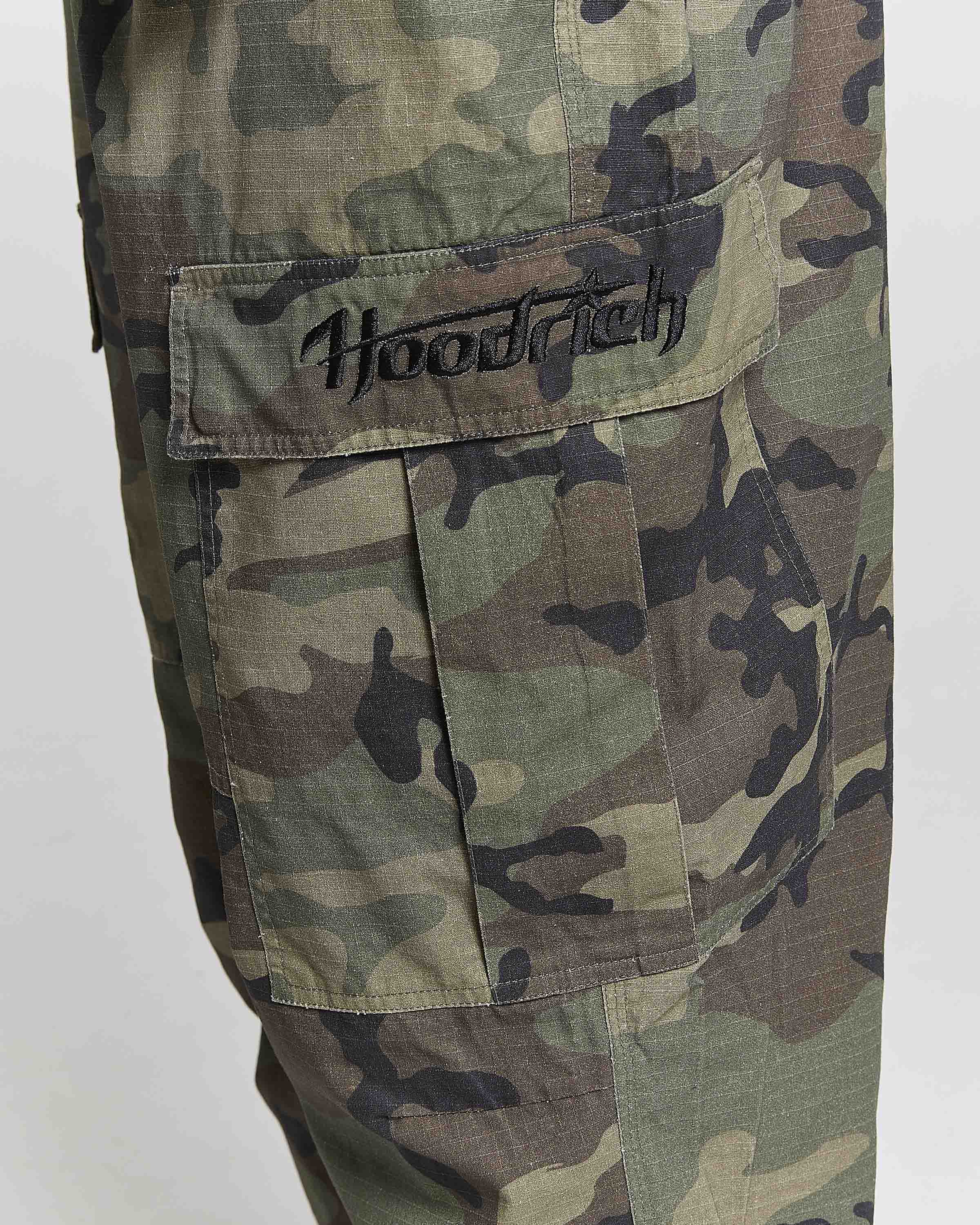Close-up of camouflage cargo pants featuring a large flap pocket with 'Hoodrich' embroidered in black script on the tab, set against a plain light gray background.
