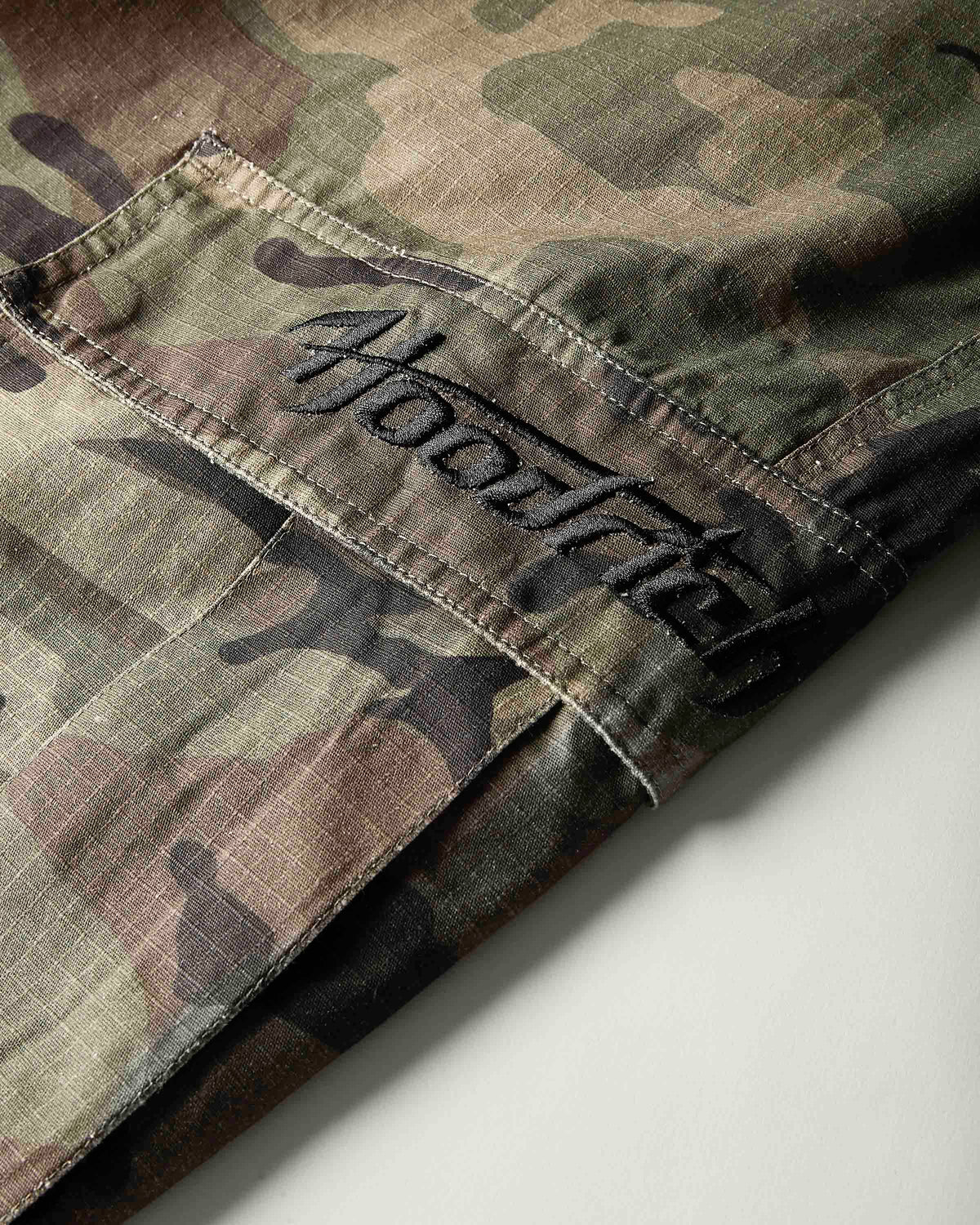 Close-up of a camouflage garment, likely a jacket or overshirt, showing a pocket with black embroidered script reading 'hoodRICH'. The fabric has a woodland camo pattern in green, brown, tan, and black.