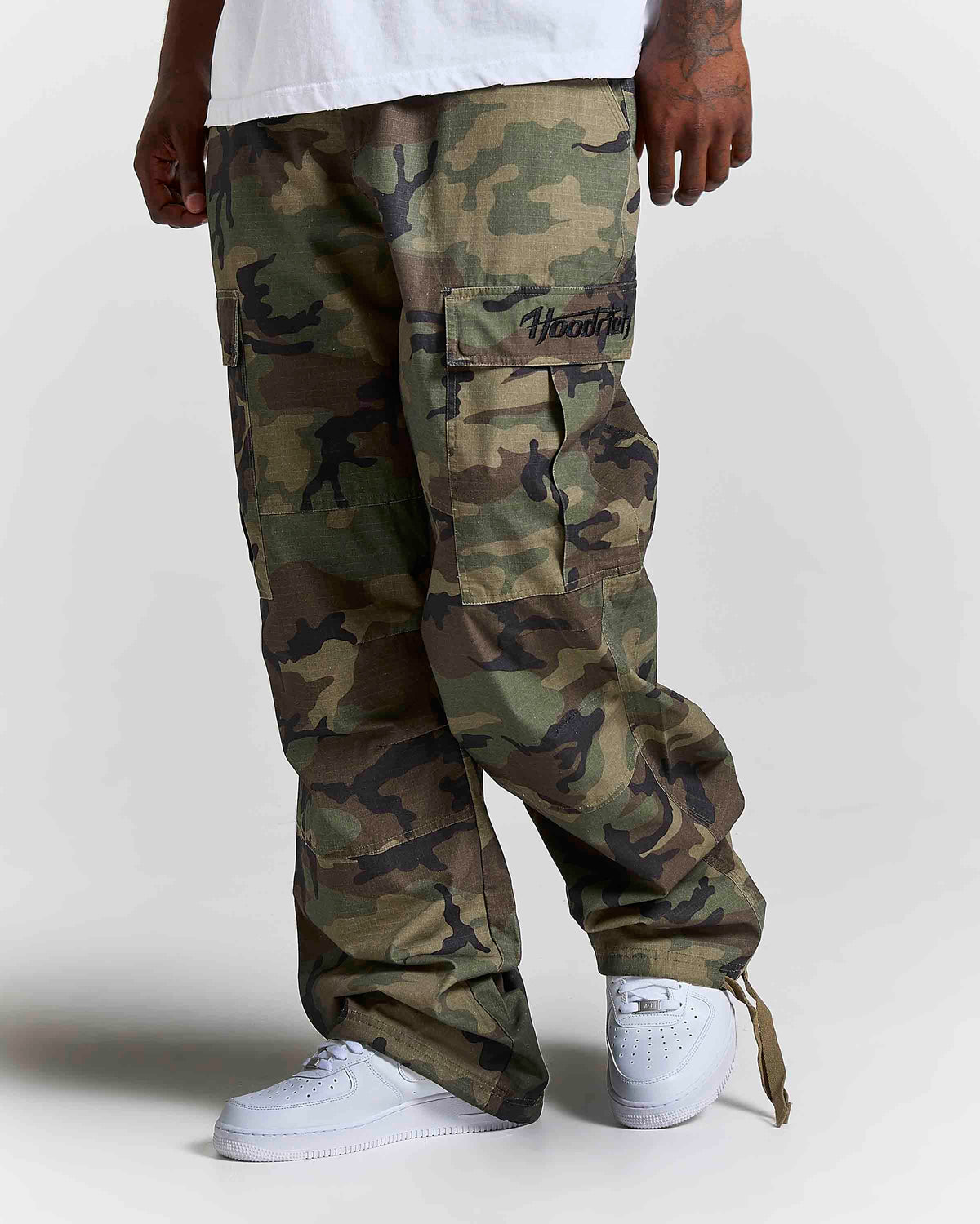 A person wearing camouflage cargo pants with a white t-shirt and white sneakers, shown from the waist down against a light gray studio background. The brand name 'HOODRICH' is embroidered on the cargo pocket.